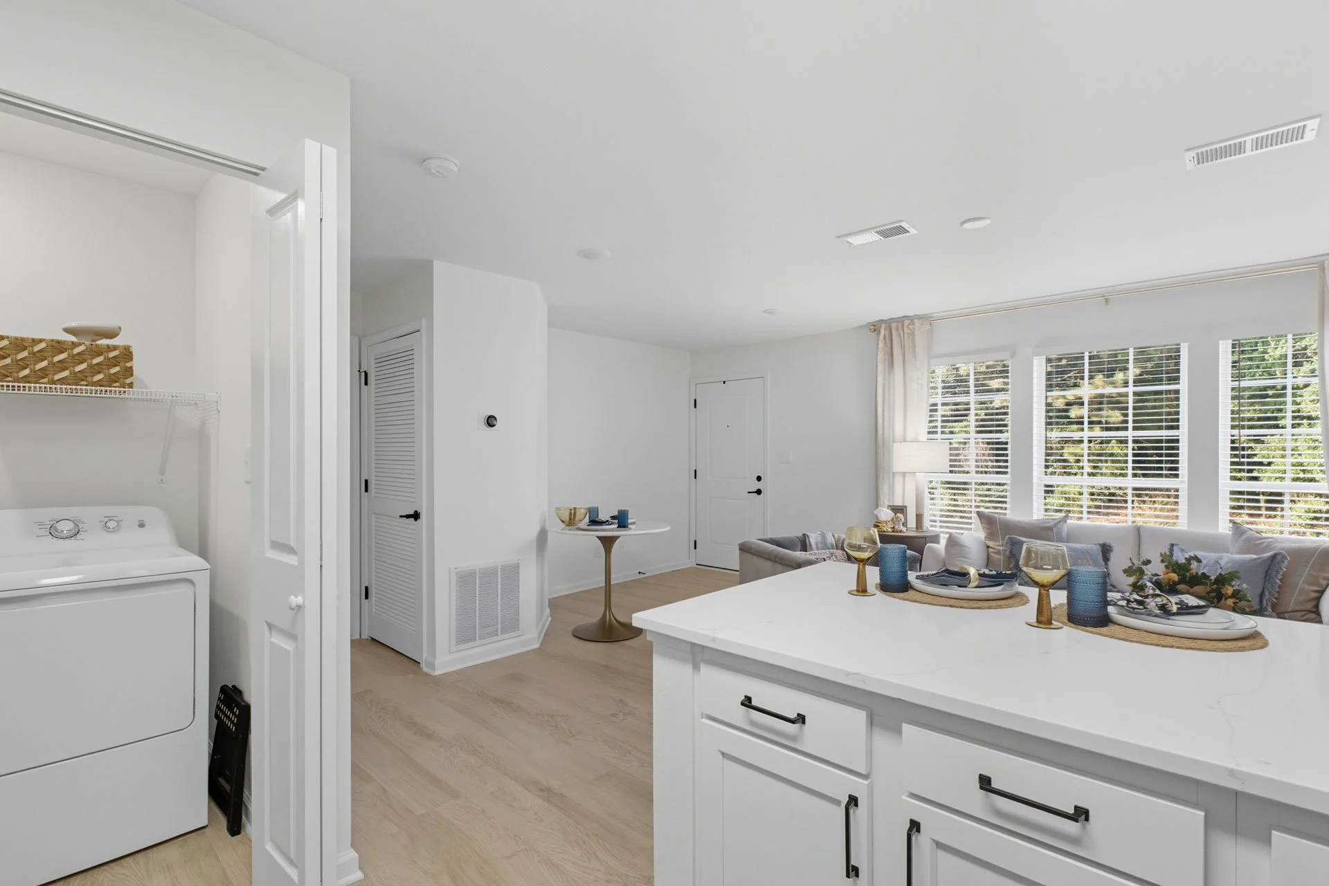 Open-concept living area with a white kitchen island, a gray sofa with throw pillows, large windows with blinds, and a small round table near the entry door.