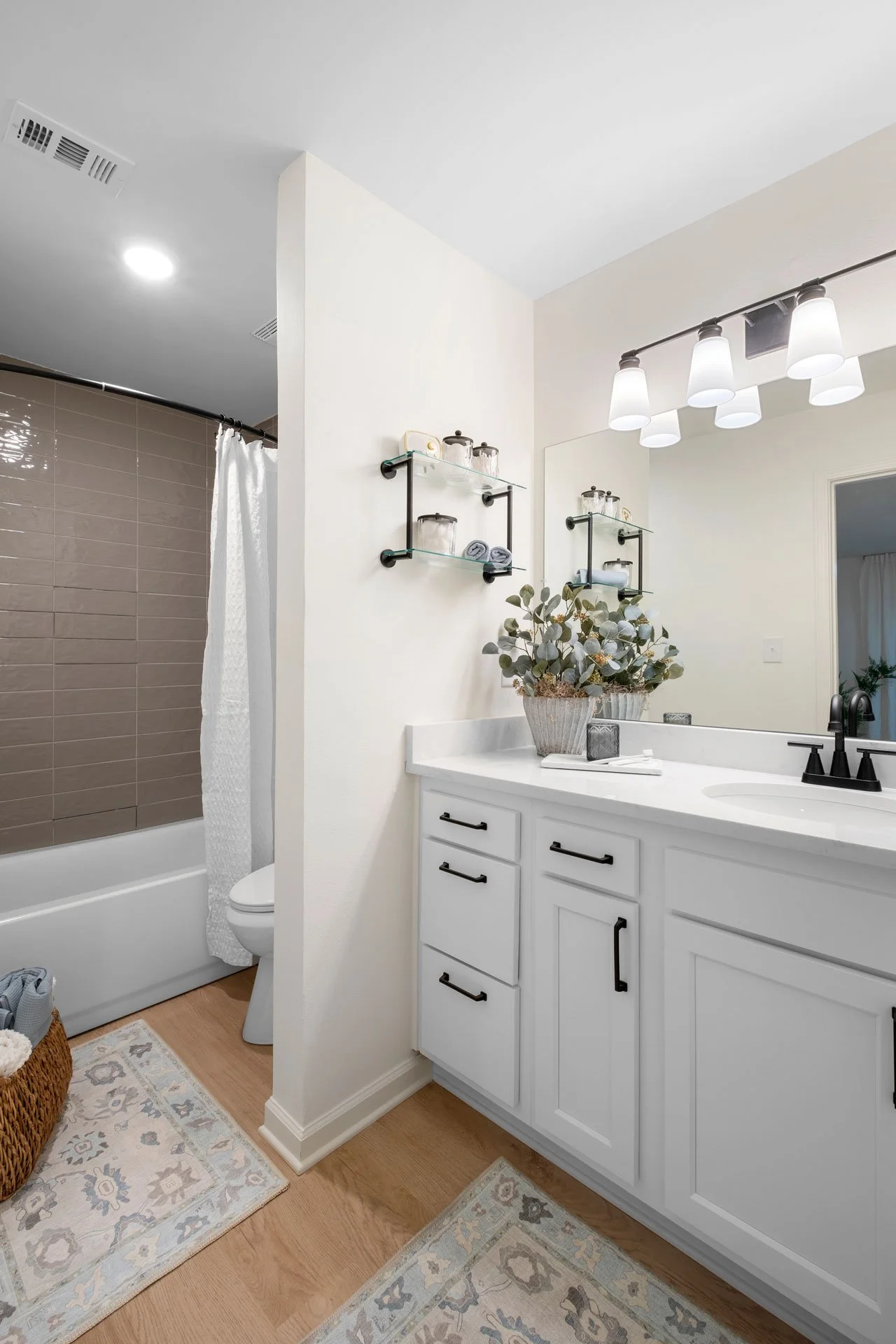 Bathroom with white vanity, large mirror, and black fixtures. Shower area with brown tiles and white curtain. Wooden floor with patterned rugs and a woven basket nearby. Decorative shelf with jars and towels, and a potted plant on the vanity.