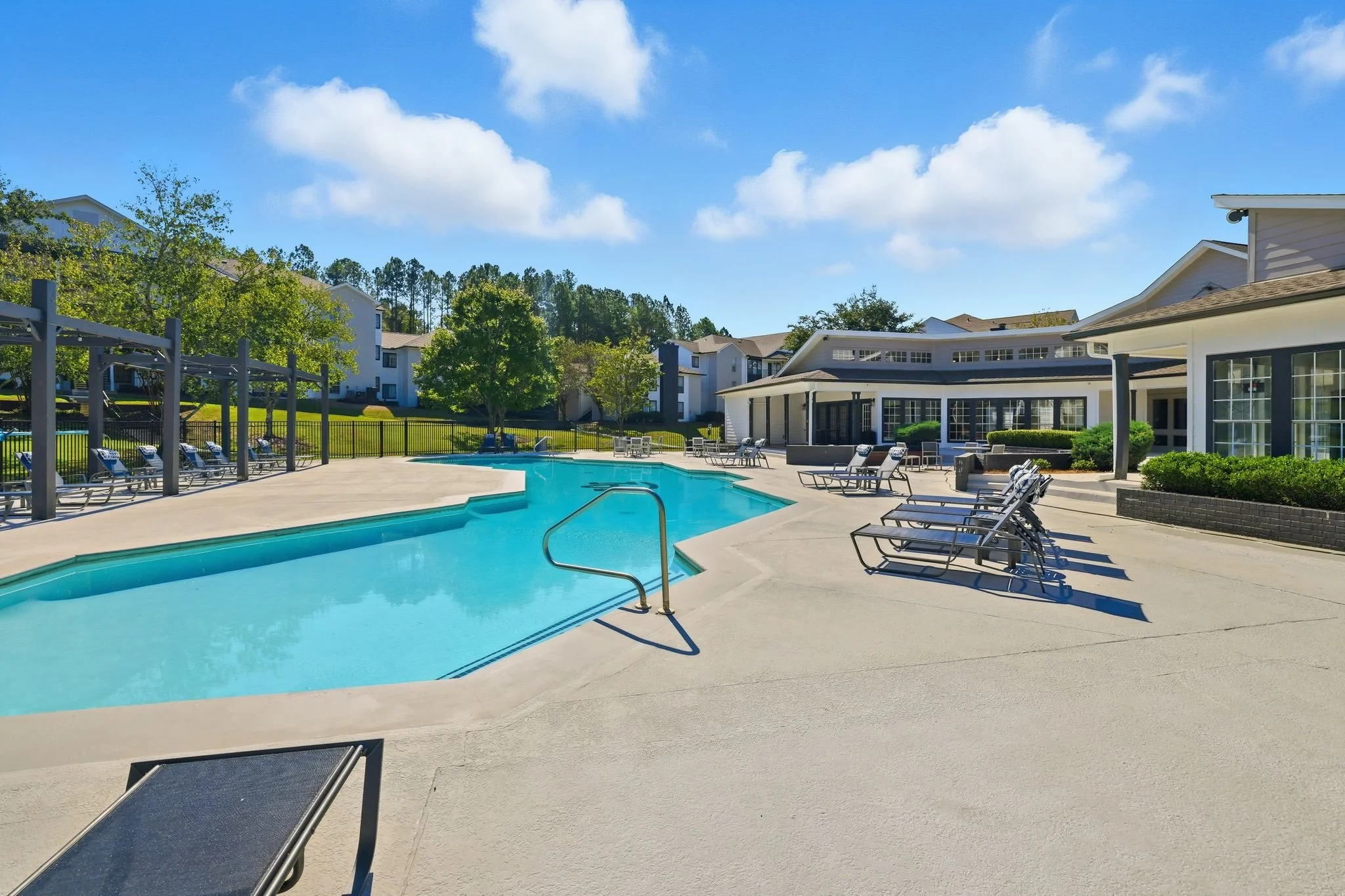 An outdoor swimming pool area with lounge chairs, a covered patio, surrounding buildings, green trees, and a partly cloudy sky.