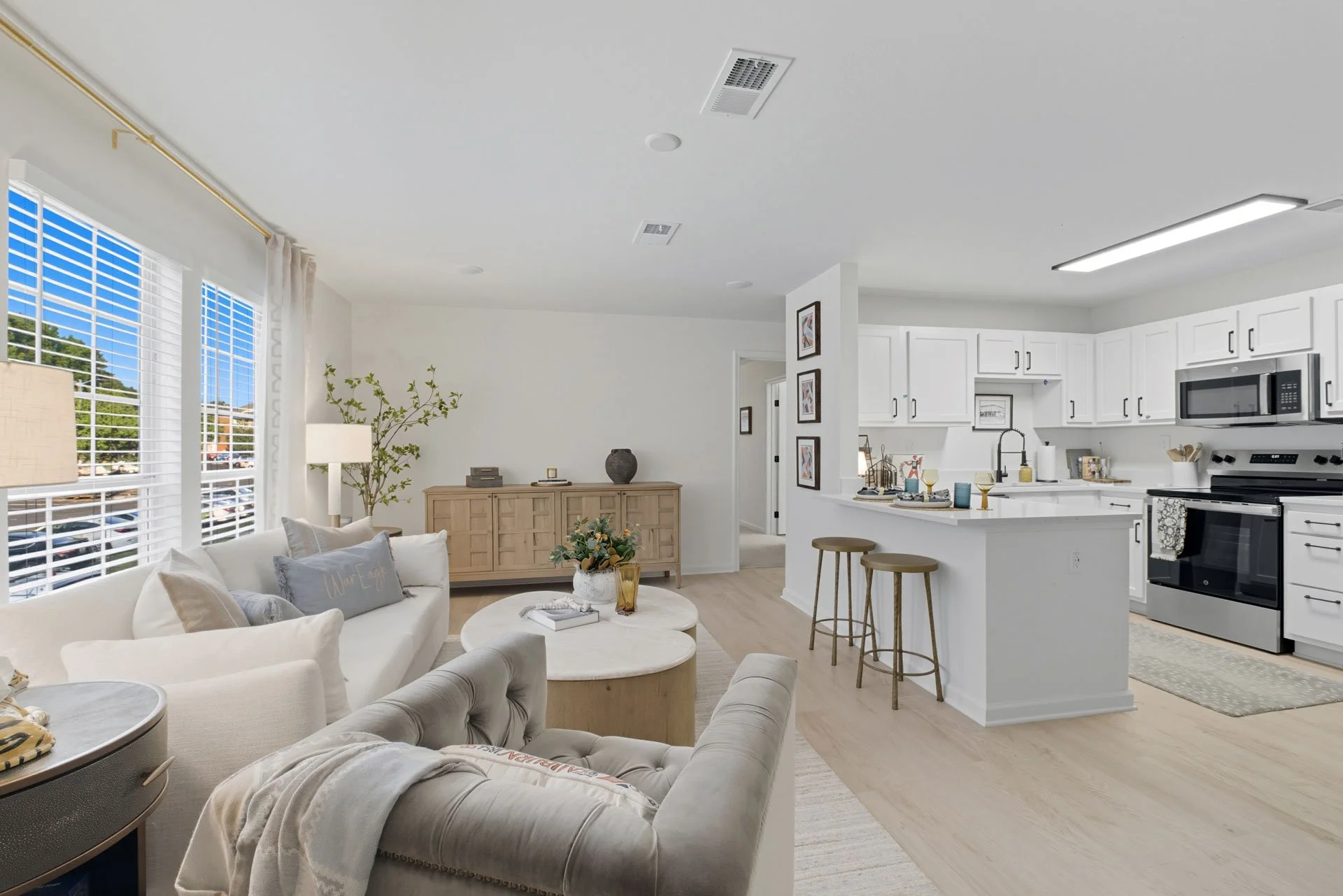 Bright open-concept kitchen and living room with white cabinets, stainless steel appliances, a breakfast bar, beige and white furniture, a large window with blinds, and decorative accents.