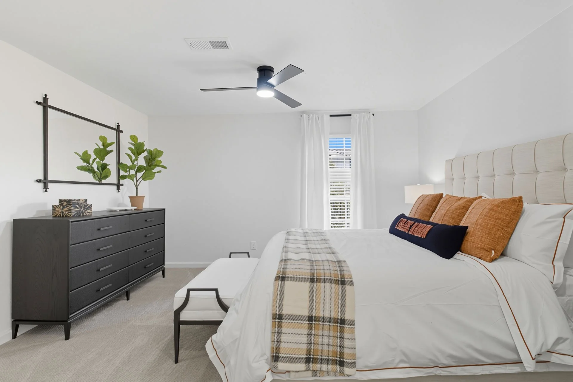 Modern bedroom with white walls, beige carpet, white bed with brown and blue pillows, plaid throw blanket, black dresser with green plant, wall mirror, and window with white curtains.