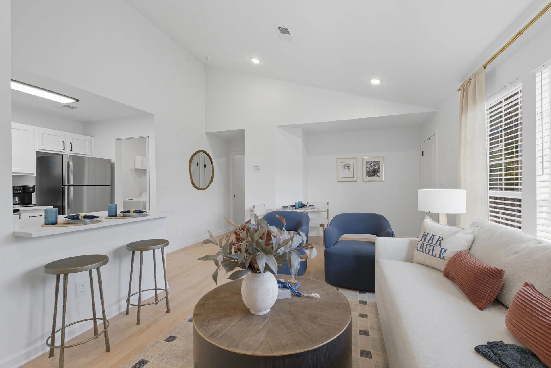 Bright living room with white walls, a beige couch with red and blue pillows, a round wooden coffee table with a white vase of leaves, two blue armchairs, a white lamp, and large windows with white blinds. Open kitchen with white cabinets, stainless 