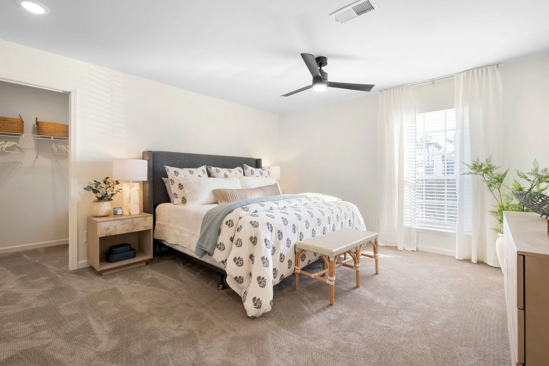 A bright, modern bedroom with a large bed, white and patterned bedding, a bedside table with a lamp, a small bench at the foot of the bed, a ceiling fan, and a window with white curtains. There is an open closet with storage baskets on the left and a