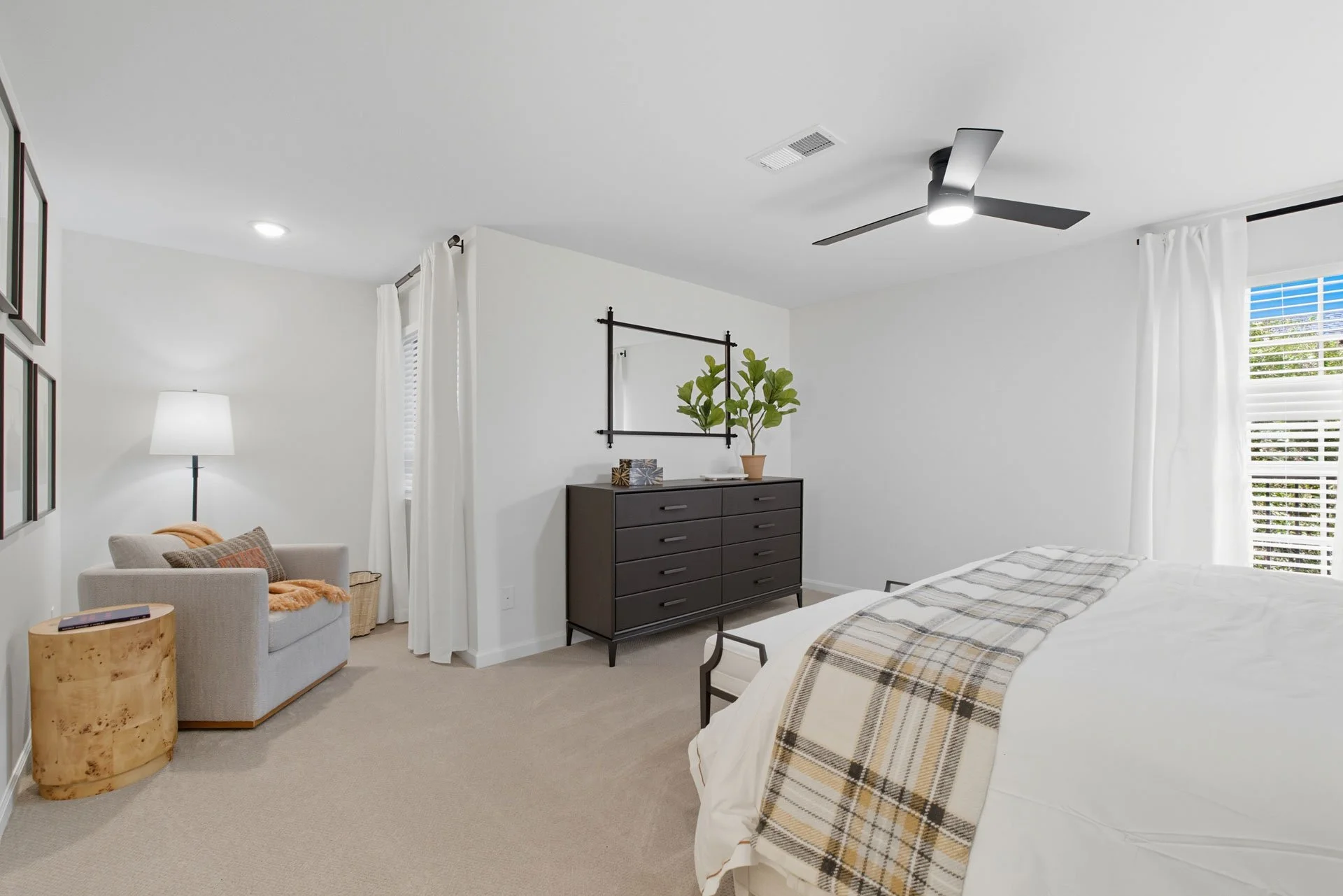 A modern bedroom with white walls, beige carpet, a bed with white sheets and plaid blanket, black dresser, gray armchair with pillows and throw, floor lamp, window with white curtains, ceiling fan, green potted plant, and decorative wall mirror.