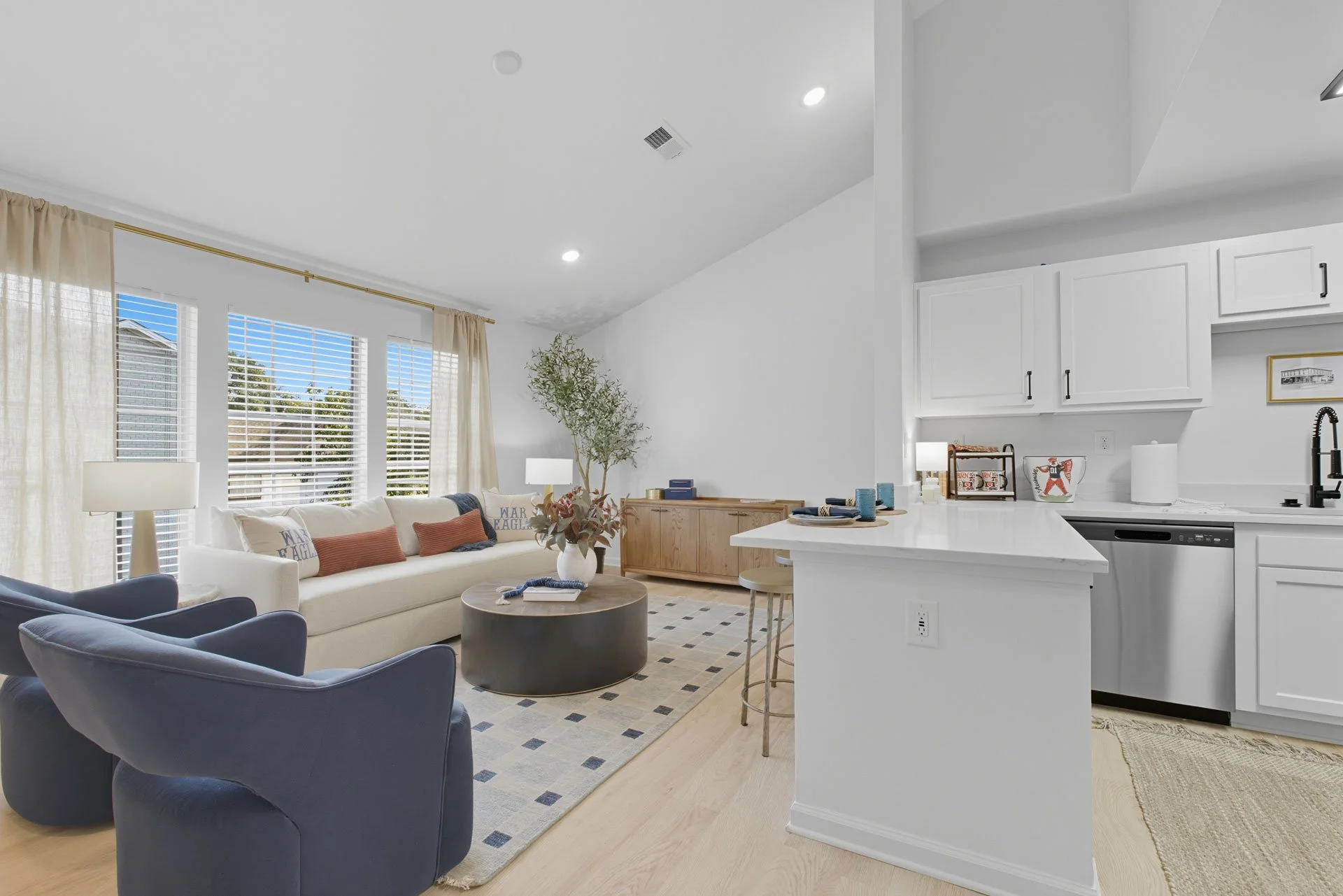 Bright living room with large windows, white walls, a white sofa with rust and navy pillows, two navy armchairs, a round coffee table with a floral vase, and cream curtains. Open kitchen with white cabinets and a black faucet.