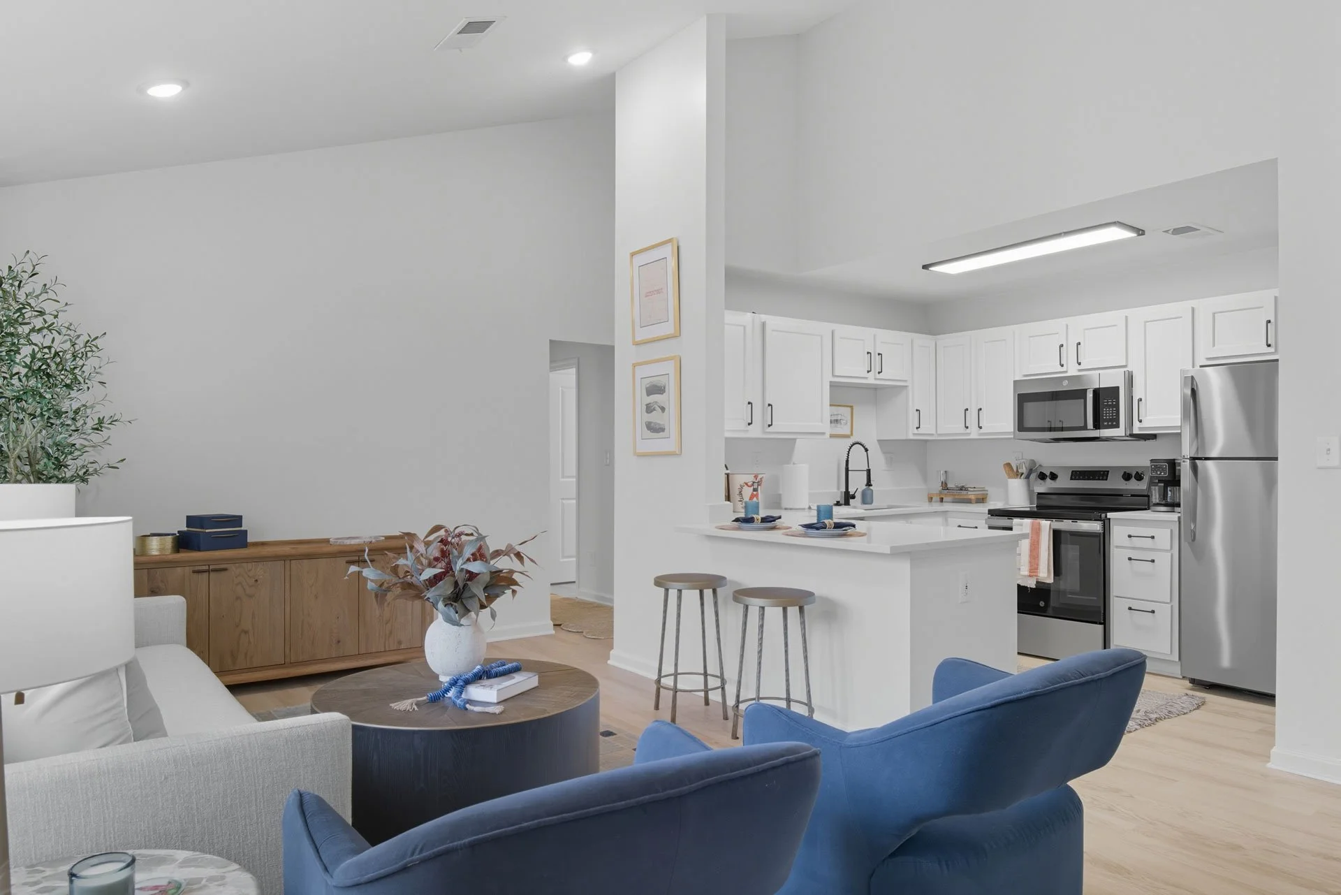 Open-concept living room with white walls, light wood flooring, a beige sofa, blue armchairs, a round coffee table with decorative items, and a wooden sideboard with books and decor. The kitchen has white cabinets, stainless steel appliances, a black