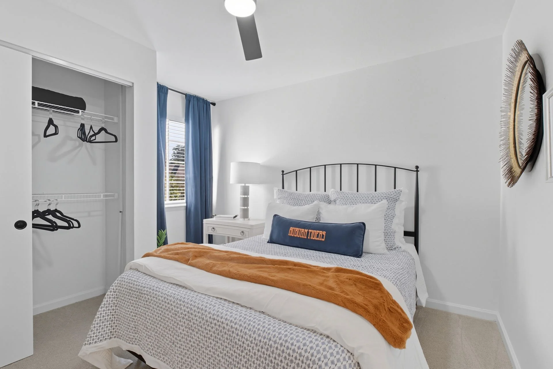 A neatly made bedroom with a bed featuring white and gray bedding, an orange throw blanket, and a navy blue decorative pillow with orange text that says "Auburn Tigers." There is a white nightstand with a lamp and a window with blue curtains beside i