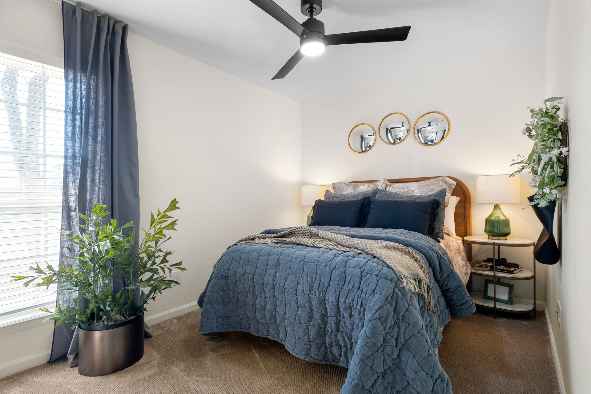 A bedroom with a bed covered in a blue quilt, multiple pillows, and a blanket. There are three round wall mirrors above the bed. On the nightstand, there's a green lamp and some books. A potted plant is next to a window with blue curtains, and a ceil