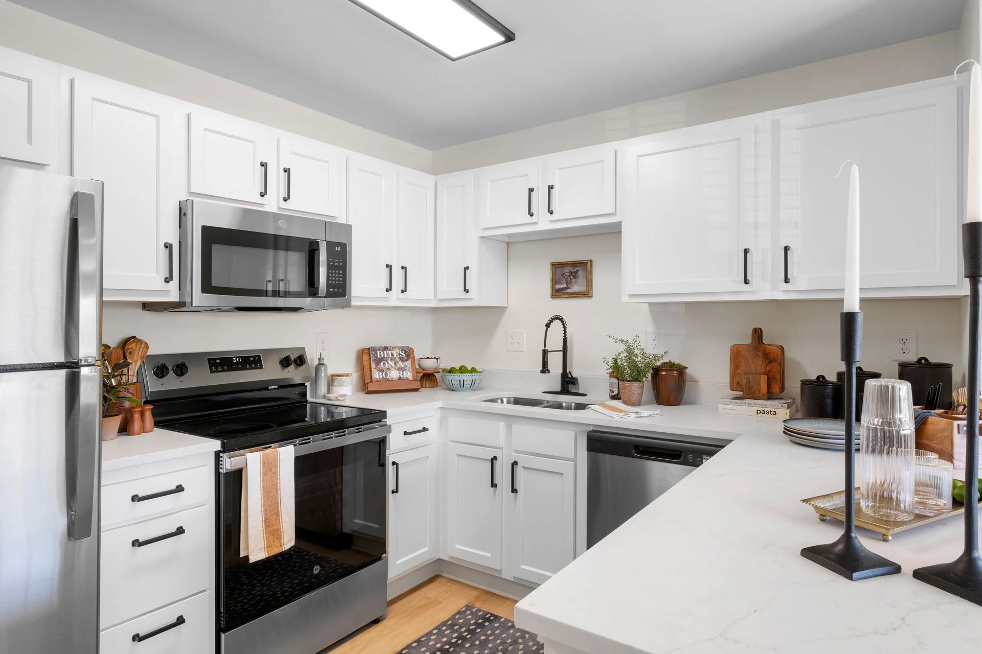 A modern kitchen with white cabinets, stainless steel appliances, a microwave, stove, dishwasher, and a refrigerator. Decor includes plants, cutting boards, and candles.