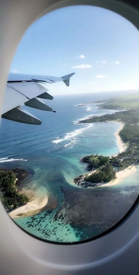 Vue d'un avion survolant une côte avec des plages, des eaux turquoise et des arbres, vue depuis une fenêtre d'avion.