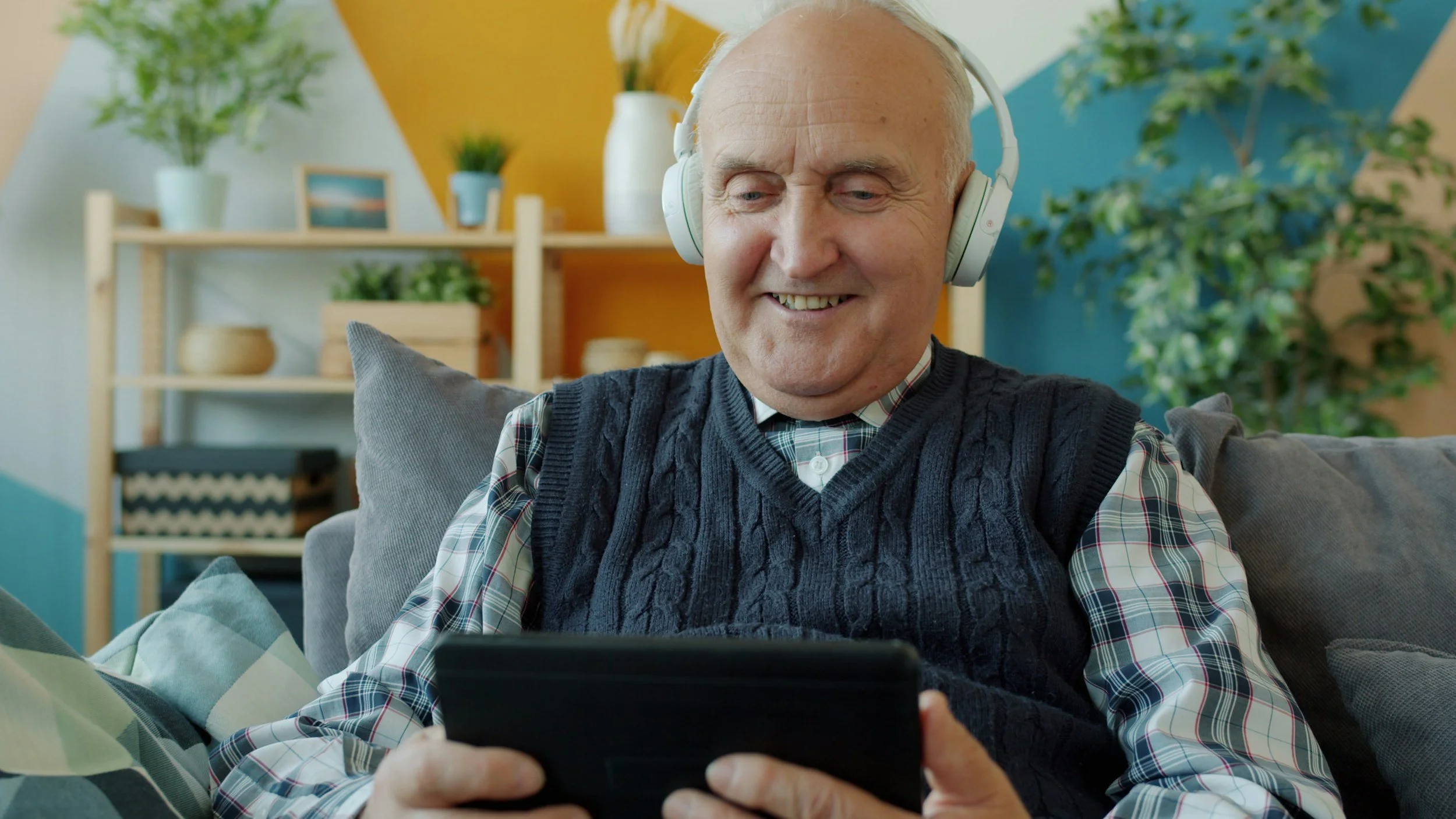 An older man in a blue sweater vest and plaid shirt sits in a chair and looks at his tablet. He has white over-ear headphones on and appears to be chucking at a movie he is watching.