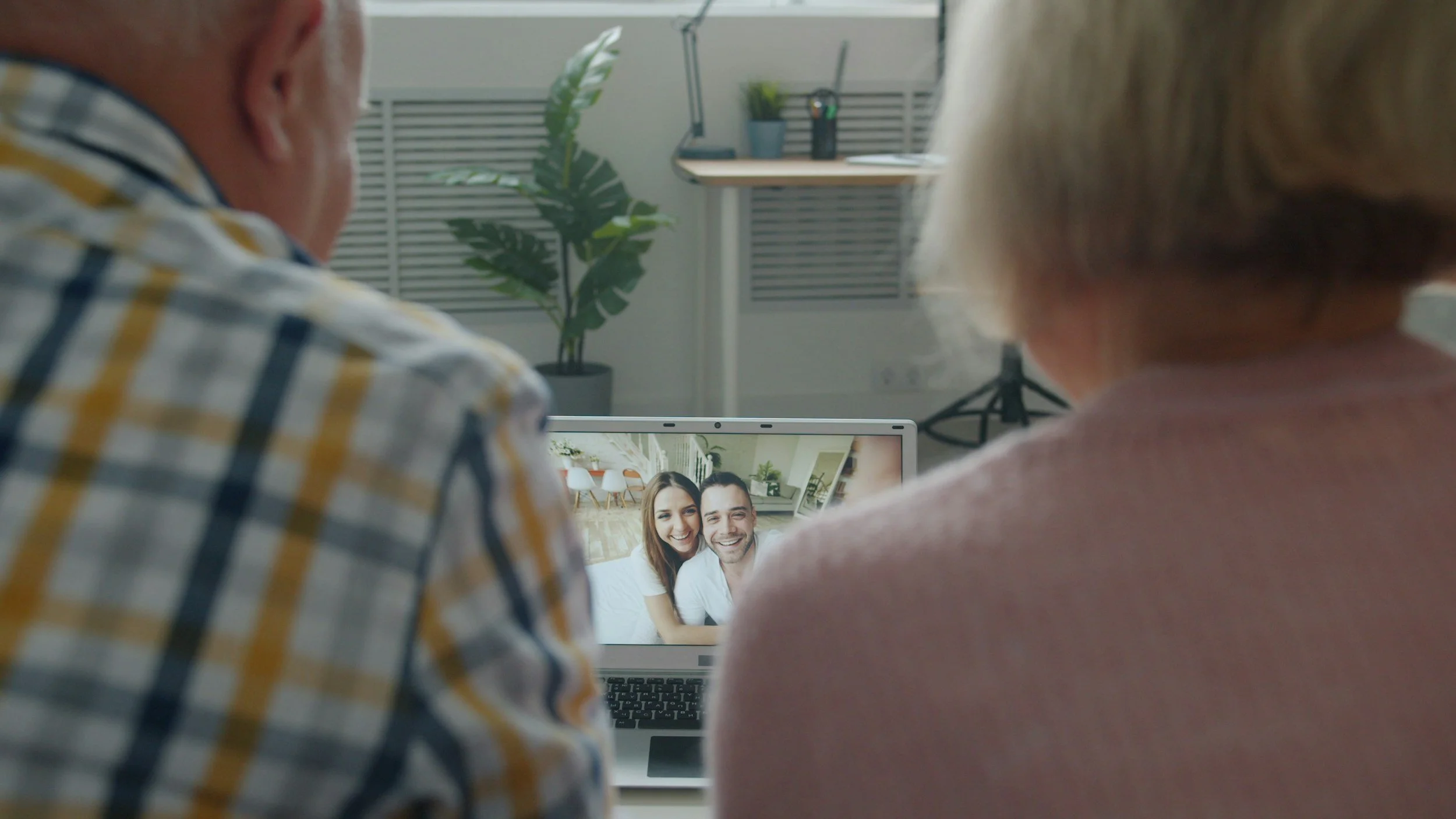 An older man and woman are seen from behind. They are looking at a laptop with an image of a smiling young man and woman on the screen who are probably their children.