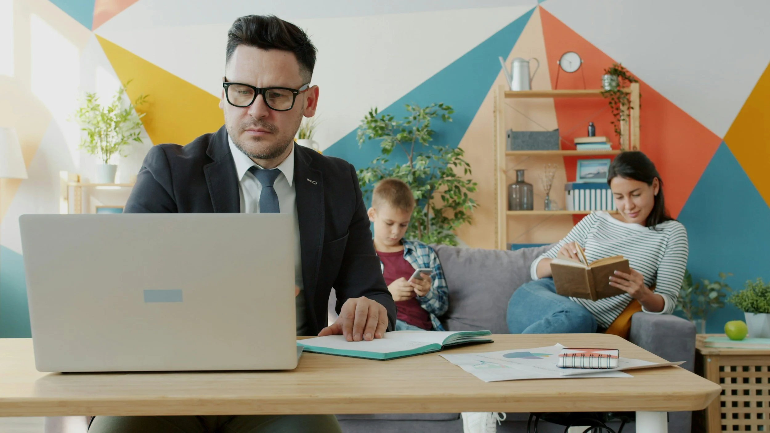 A man in a suit and a blue tie sits at a table in front of a laptop computer. His wife and son are sitting on the couch in the background. The wife is reading a book and the son is looking at a smart phone. The room is painted in bright colors.