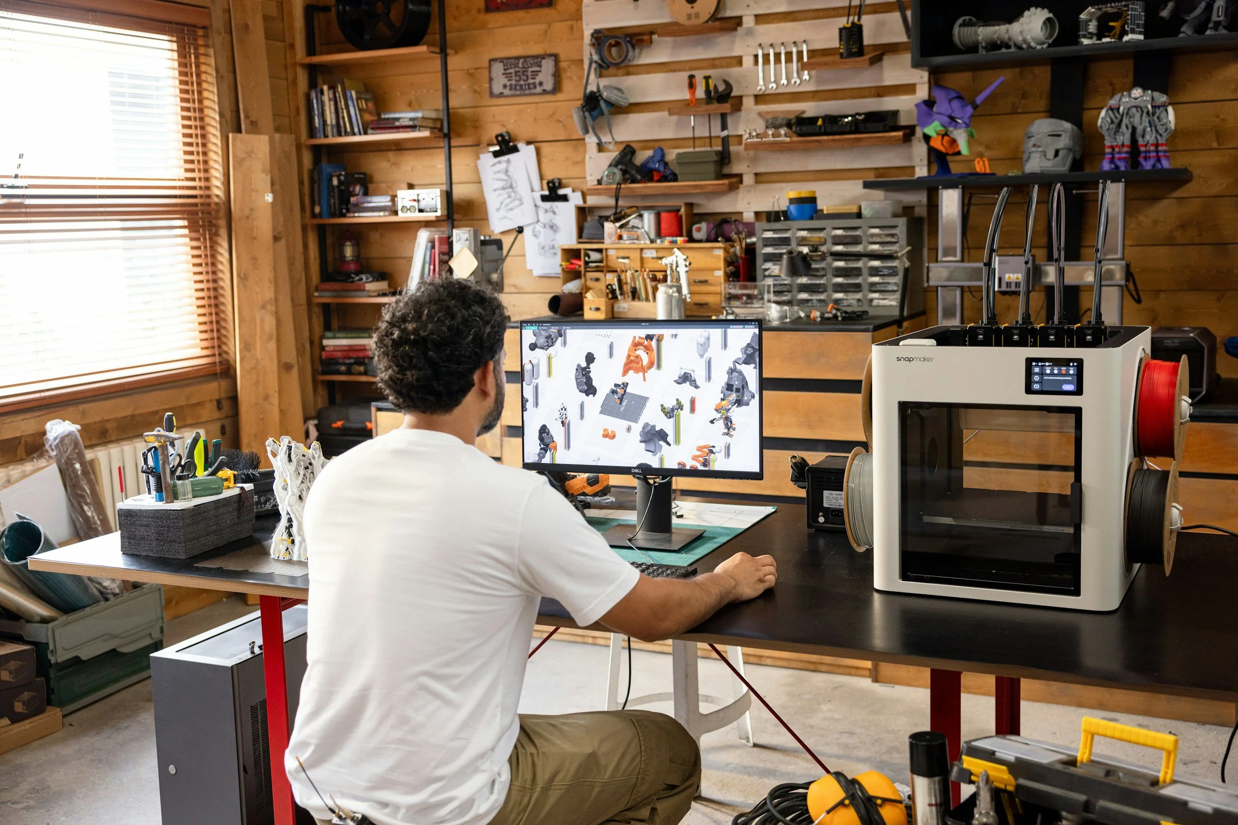 A man in a white t-shirt works on designs on his computer. Next to his computer is a 3D printer. He appears to be in a workshop surrounded by lots of tools.