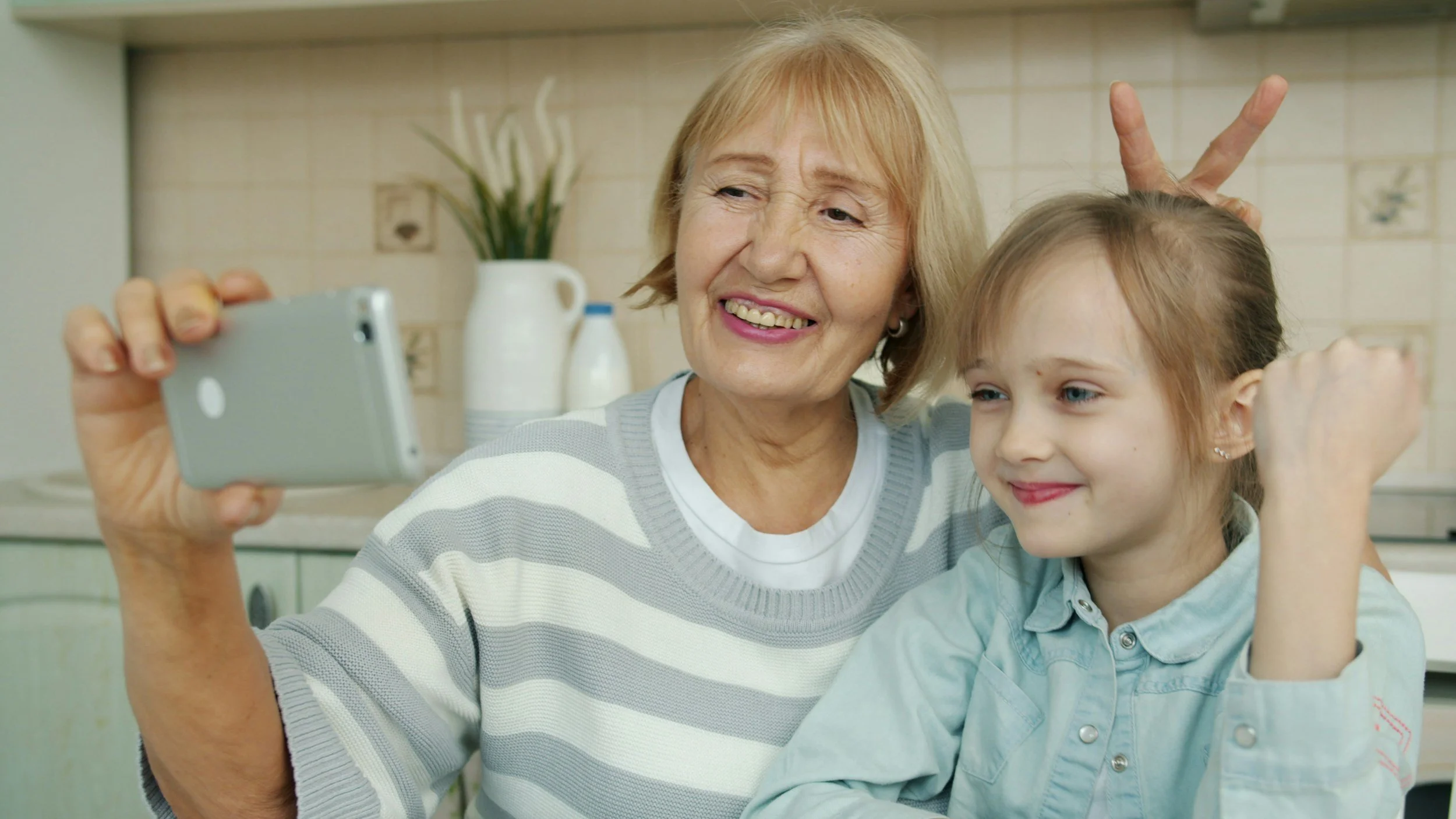 A grandma holds out her smart phone to take a selfie with her granddaughter. They are both smiling. The grandma holds "bunny ears" up behind the granddaughter's head.