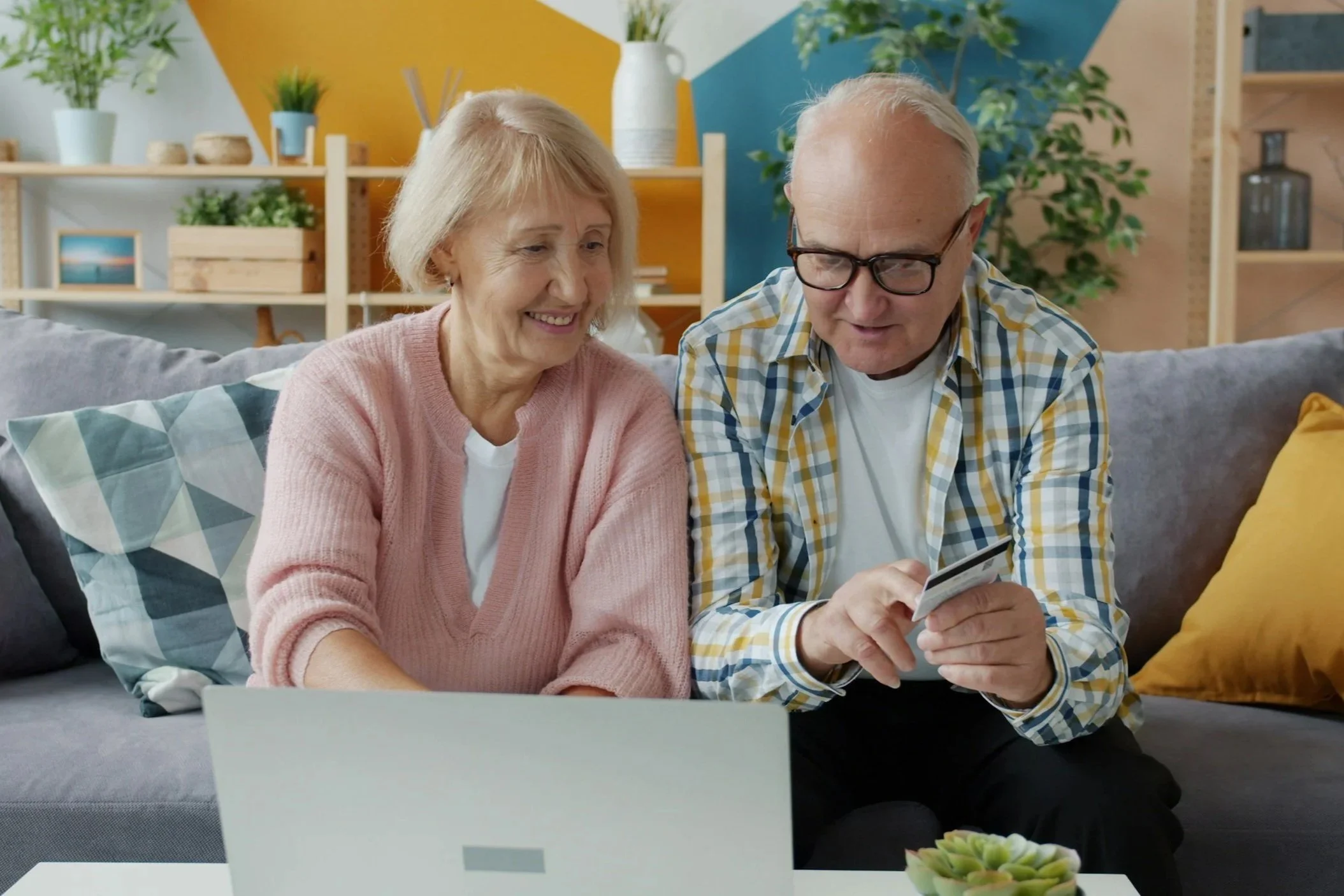 An older man and a woman sit next to each other on a couch with a computer on the coffee table in front of them. They appear to be trying to figure out how to enter payment information into a website. The senior citizens are happy.