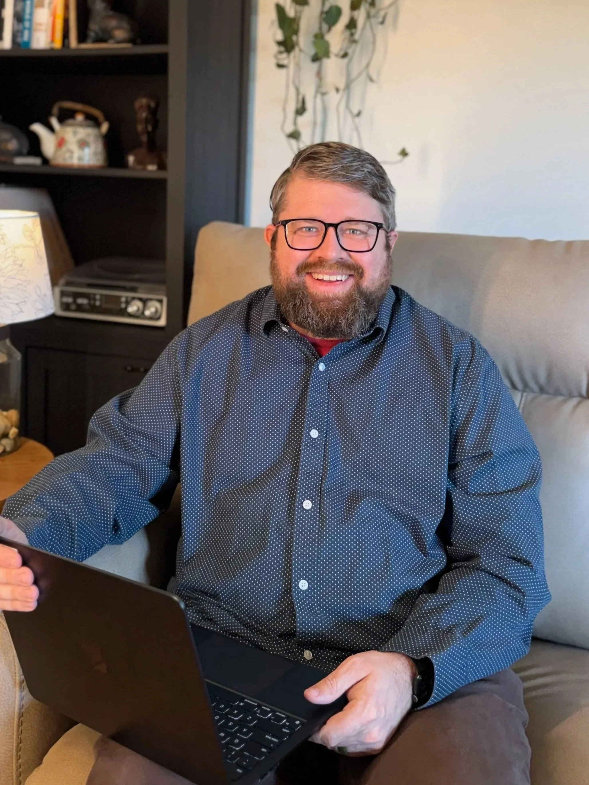 A bearded man with glasses smiles at the camera while holding a laptop in his lap.