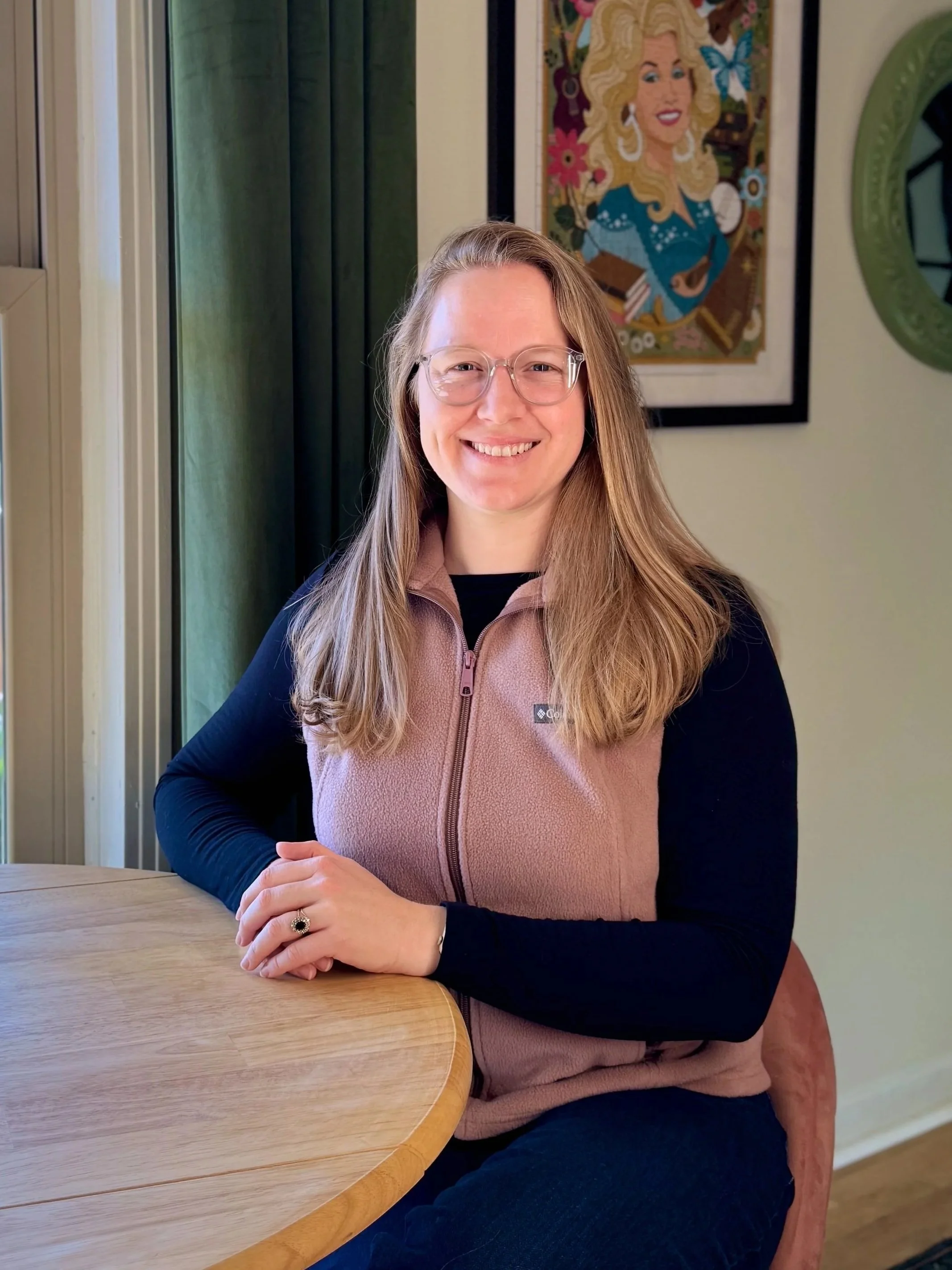 A blonde woman sits at a table and smiles at the camera. A framed puzzle of Dolly Parton hangs on the wall behind her.