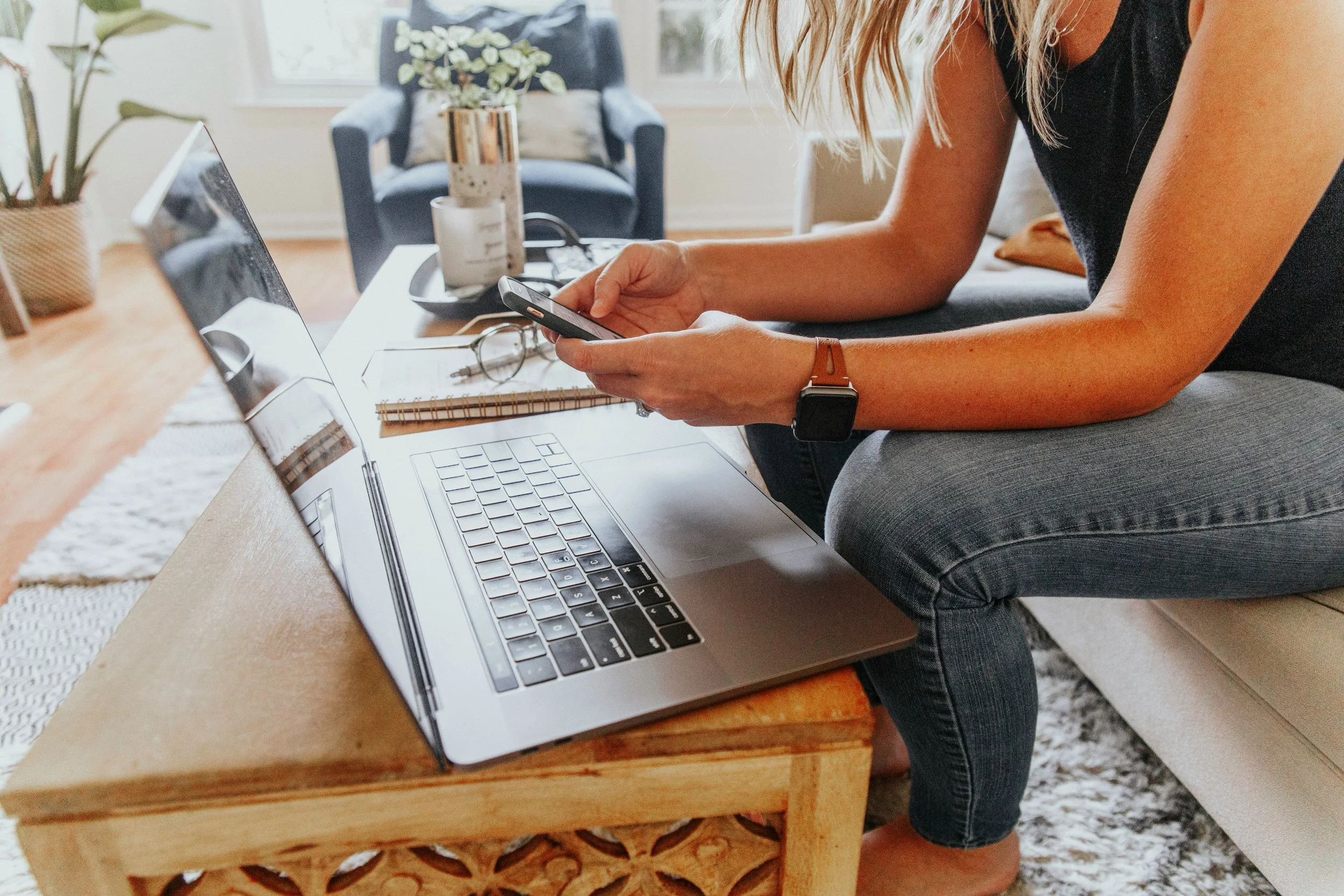 A woman sits at her coffee table with her laptop computer and looks at her smartphone. She is wearing a black tank top, jeans, and a smart watch. A notebook and a pair of glasses are on the coffee table.