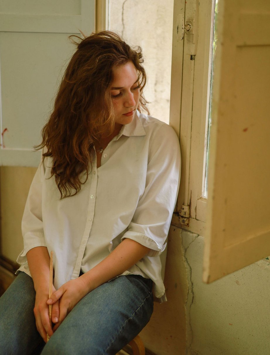 A young woman with curly brown hair, wearing a loose white button-up shirt and jeans, sitting by a window with sunlight coming in, looking down pensively.