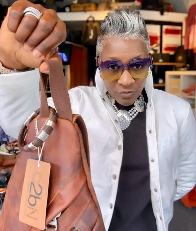 A person with short gray hair, wearing purple and yellow sunglasses, a white jacket over a black shirt, and a pearl necklace, holding up a brown purse with a tag, in a retail store setting.
