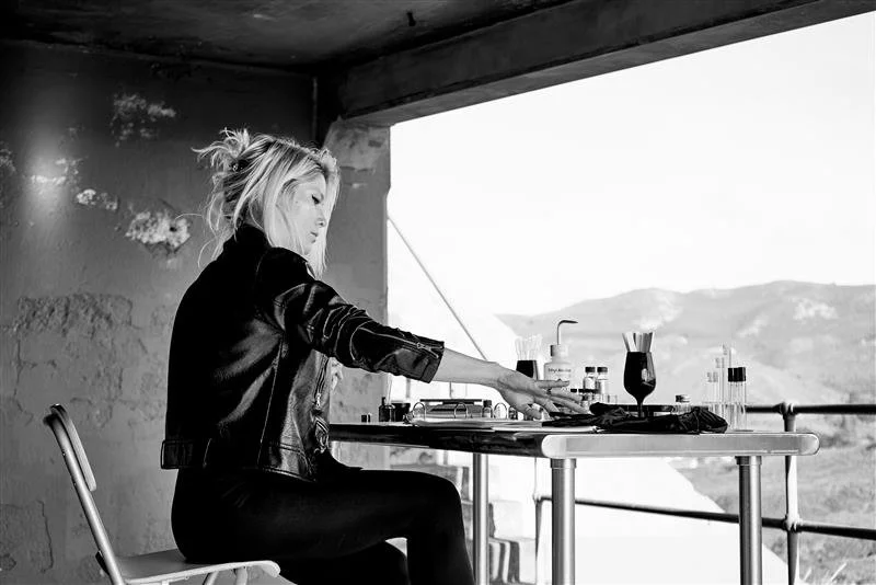 A woman reaching across a table with drinks and utensils, sitting in front of a large window showing a landscape of hills.