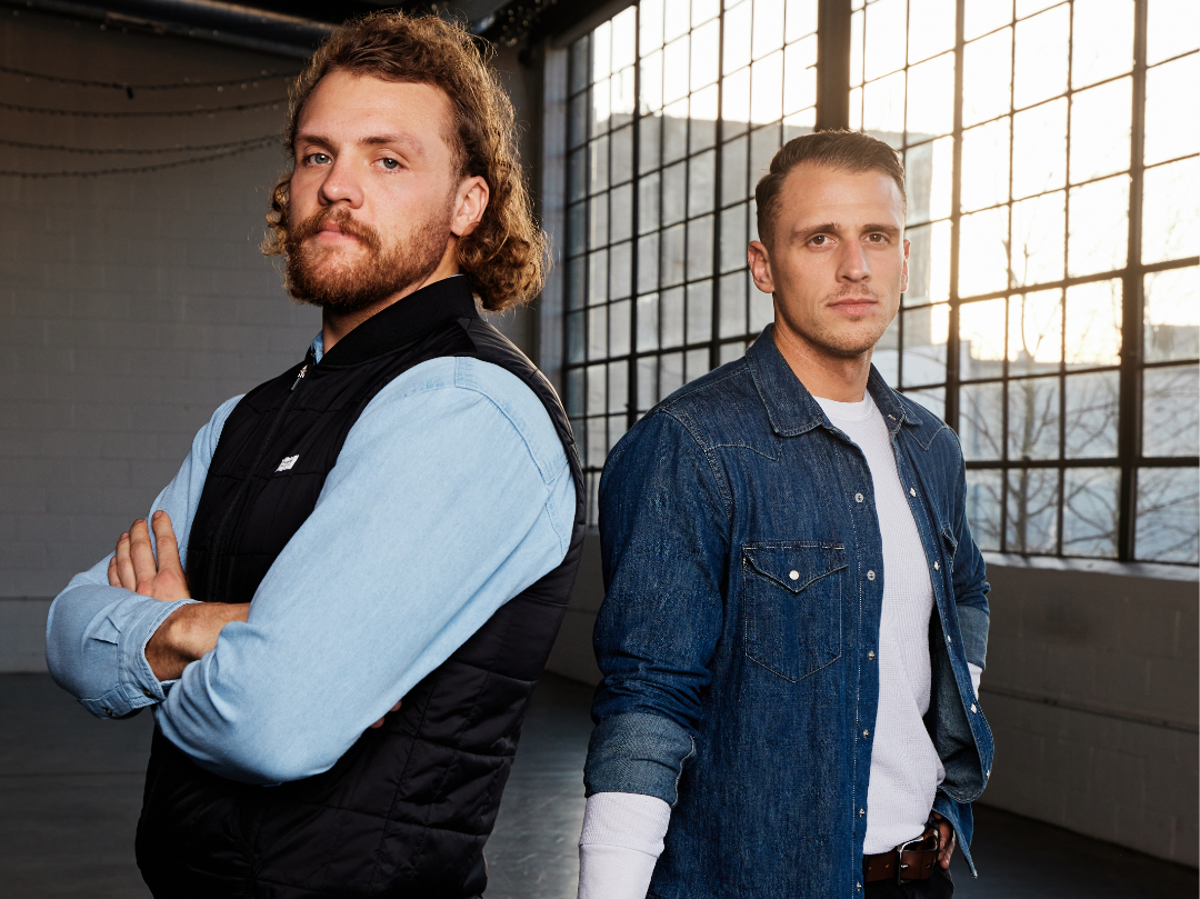 Two young men stand in a spacious industrial-style room with large grid windows and natural light, one with curly hair and a beard, wearing a black vest over a blue shirt, and the other with short hair, wearing a denim jacket and white t-shirt, both looking at the camera with serious expressions.