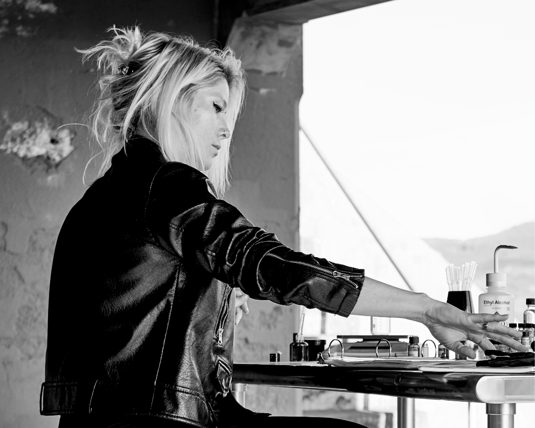 A woman with blonde hair in a messy bun wearing a black leather jacket, reaching forward with her right arm, in a room with large windows and various bottles including alcohol and syringes on a table.