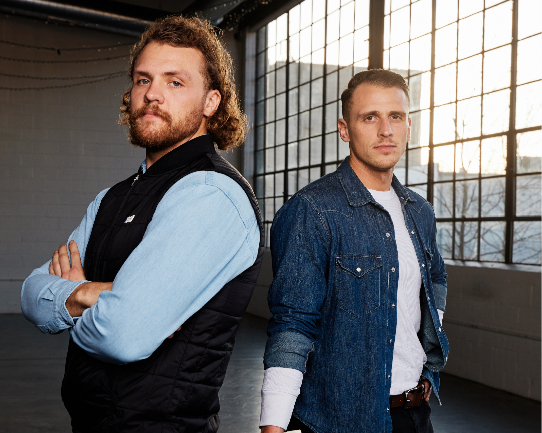 Two men standing with arms crossed in front of large windows in an industrial-style space, backlit by natural sunset light.