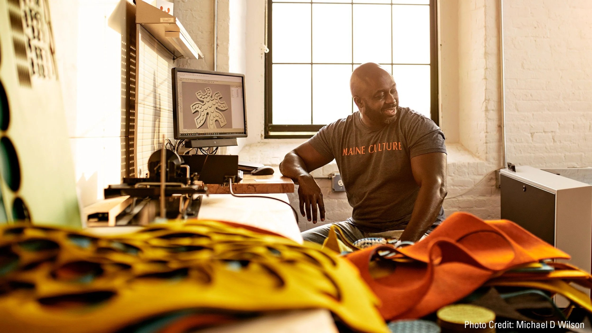 A man sitting at a desk in a creative work space, with a computer monitor displaying a stylized design, surrounded by colorful fabric or materials, in a room with a large window and white brick walls.