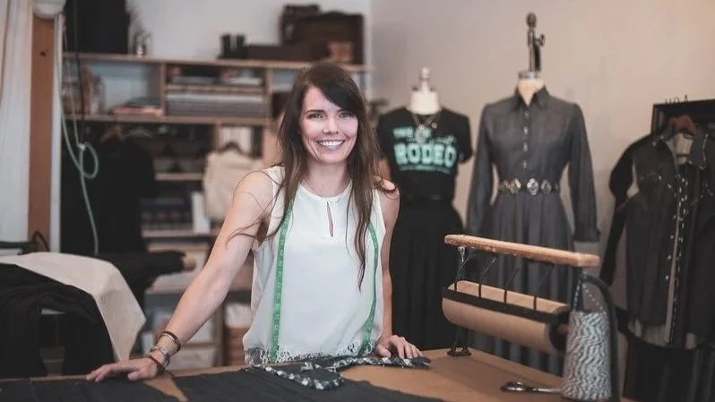 A woman standing behind a sewing table in a sewing studio, smiling, with dress forms and clothing patterns in the background.