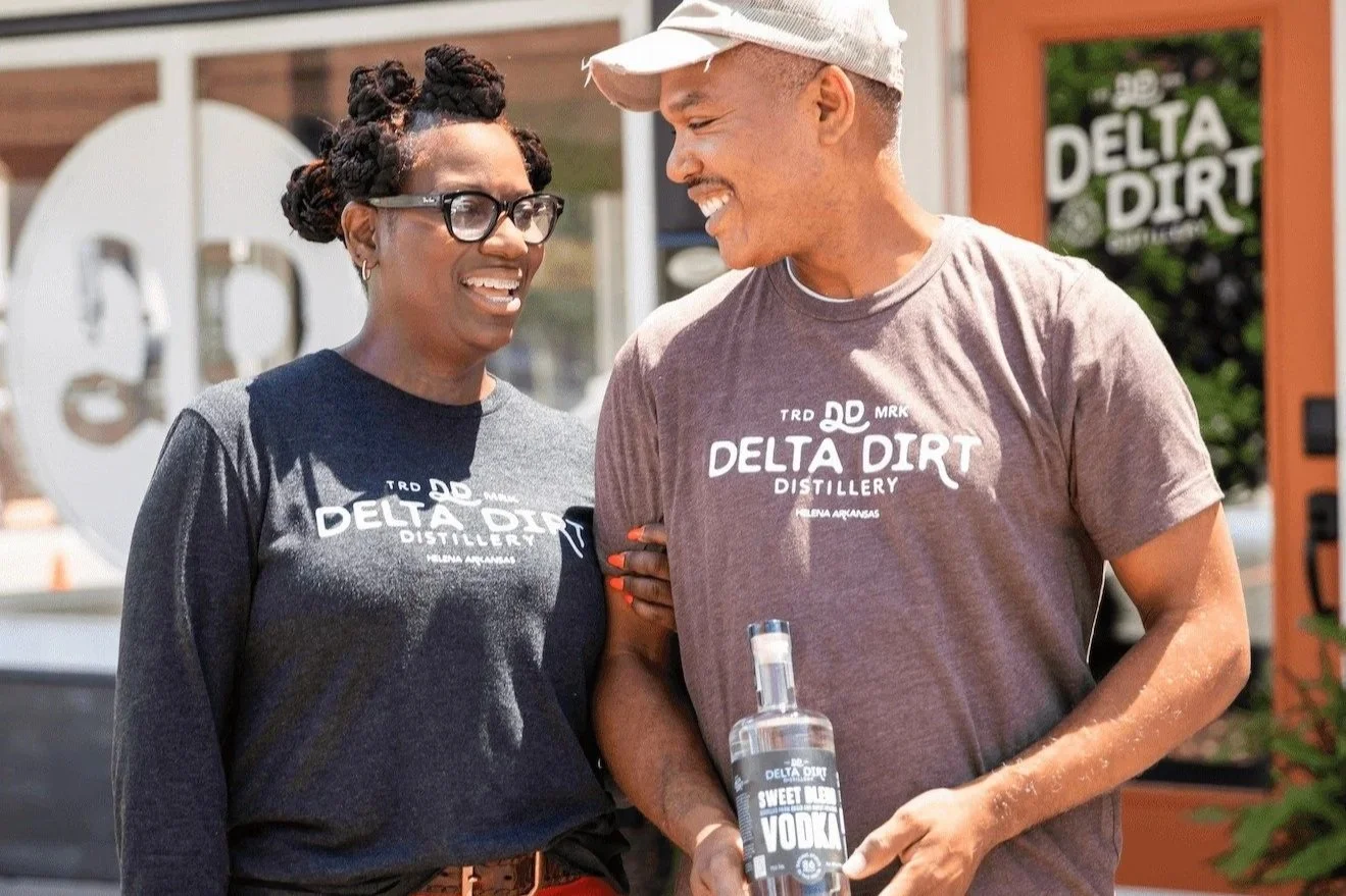 Man and woman smile and look at each other while man holds bottle reading, "Delta Dirt Sweet Blend Vodka." Owners of Delta Dirt Distillery in Helena, Arkansas.