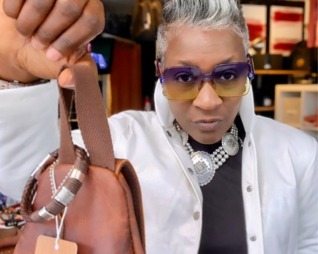 A woman with short gray hair wearing large sunglasses, a white jacket, and a black top with jewelry, holding a small brown handbag in front of her.