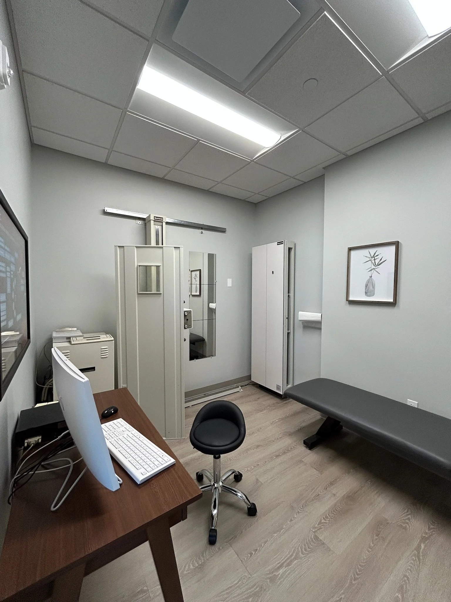Medical examination room with a patient bed, a computer desk with a monitor, keyboard, and mouse, a chair, and medical equipment including a tall gray cabinet and an X-ray viewing box on the wall. There is a framed artwork of a plant on the wall.