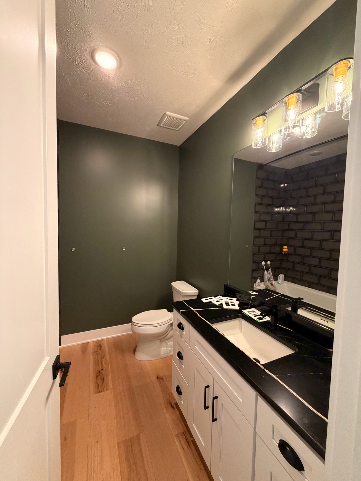 A bathroom with a white sink vanity, black countertop, and a large mirror. The wall behind the sink is dark green, and there is a brick accent wall in the shower area. The bathroom has wooden flooring, a toilet, and a black door with a handle on the 