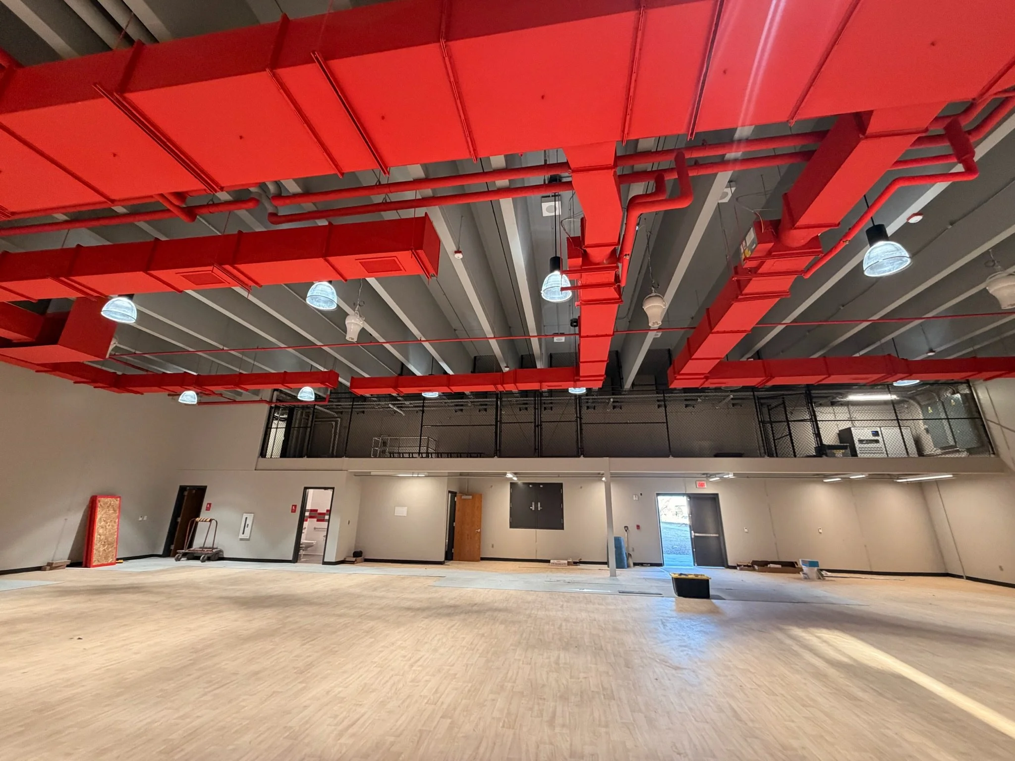 Empty room with beige flooring, gray walls, and exposed red piping on the ceiling, with some hanging lights and doors.