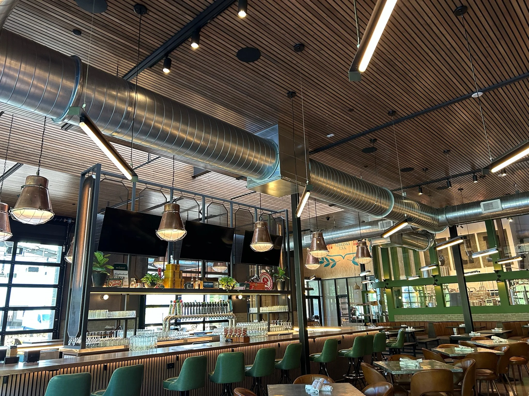 Interior of a modern restaurant or bar with a wooden slat ceiling, large metal air ducts, hanging light fixtures, and green chairs around tables.