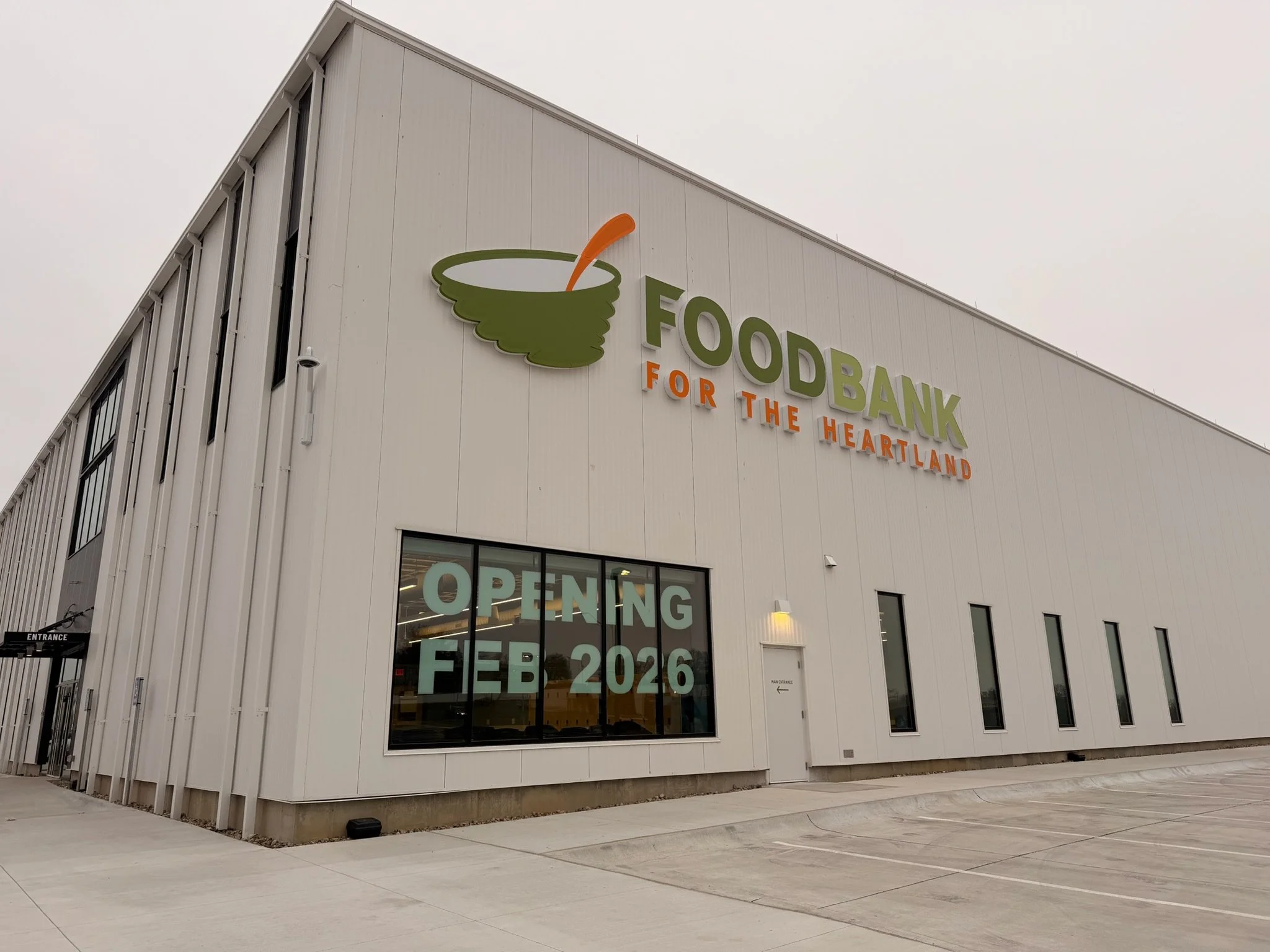 Exterior view of a modern white building with signage indicating a food bank opening in February 2026 and featuring a green and orange logo.