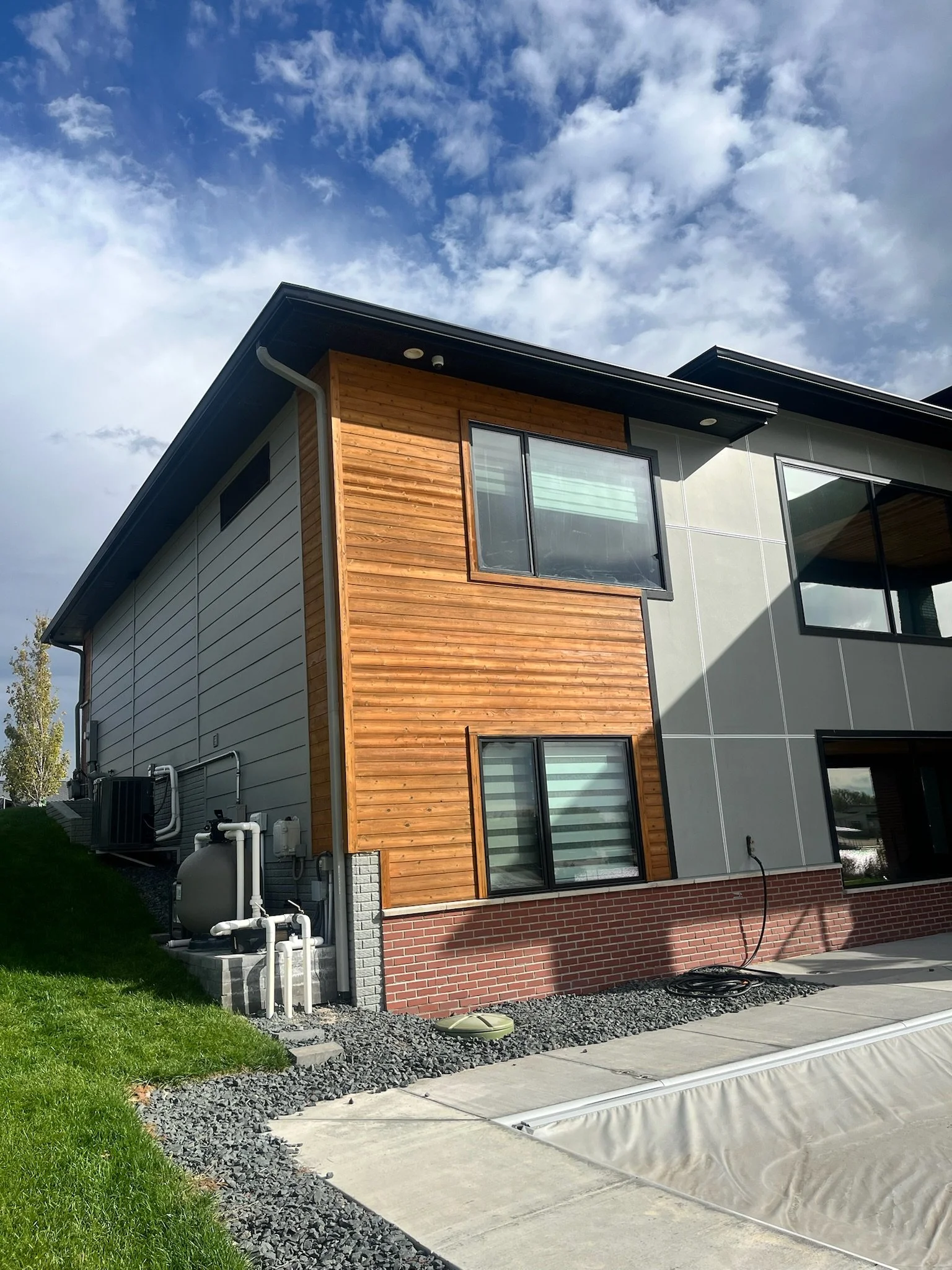 A modern house with a combination of wooden and gray exterior walls, large windows, and a brick foundation. There's a small green lawn, a concrete walkway, and some pond liner or protective cover on the ground near the house. Utilities and pipes are 