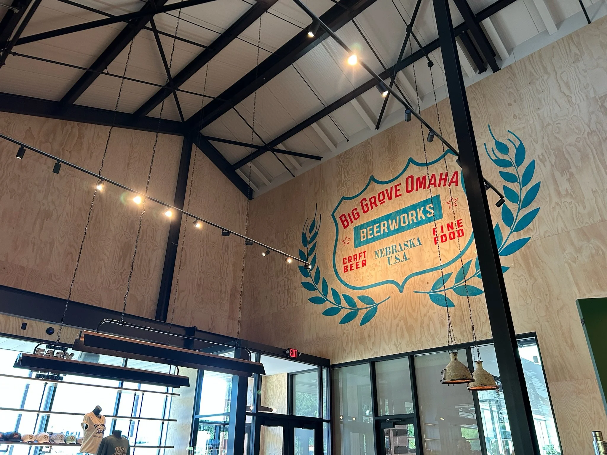 Interior view of a modern commercial space with high ceilings, exposed black metal beams, hanging pendant lights, and a branded wall sign reading "Big Grove Omaha Brew Works" with decorative blue laurel branches, large windows, and merchandise displa