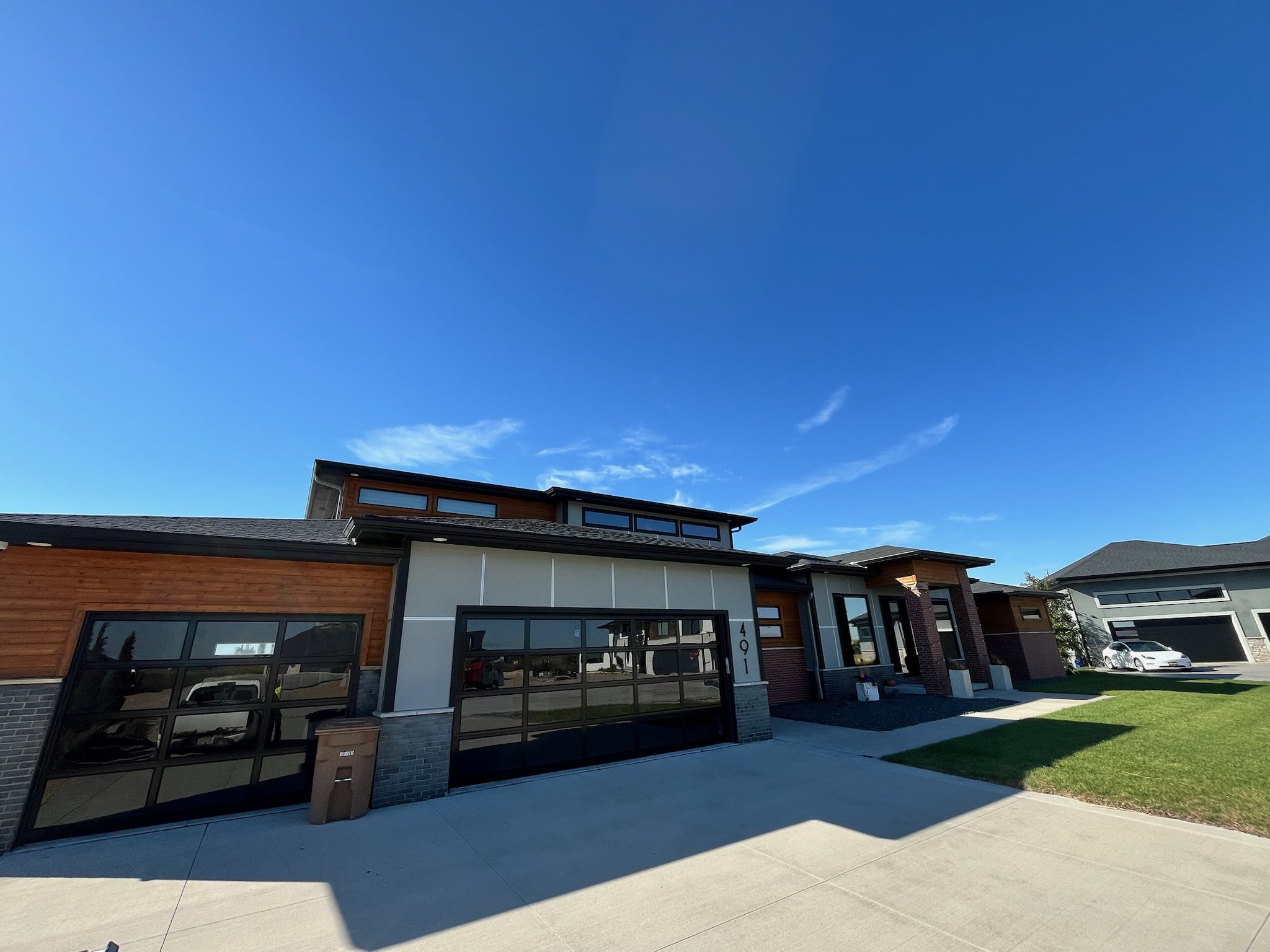 A modern residential building with a large glass garage door, a small grassy lawn, a sidewalk, and cars parked outside under a clear blue sky.