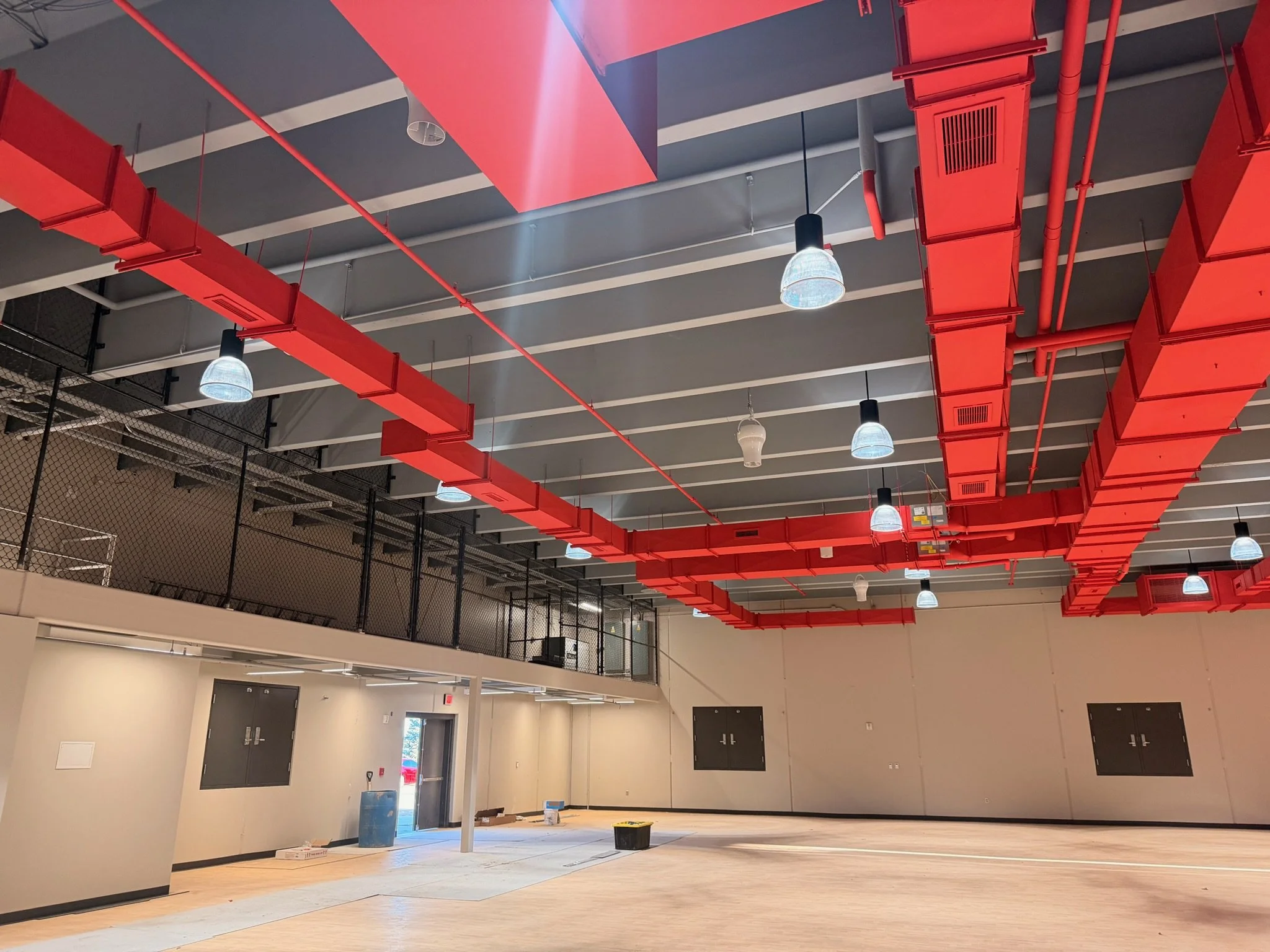 Interior of a building under renovation with a high ceiling, red ductwork, hanging light fixtures, and construction materials on the floor.