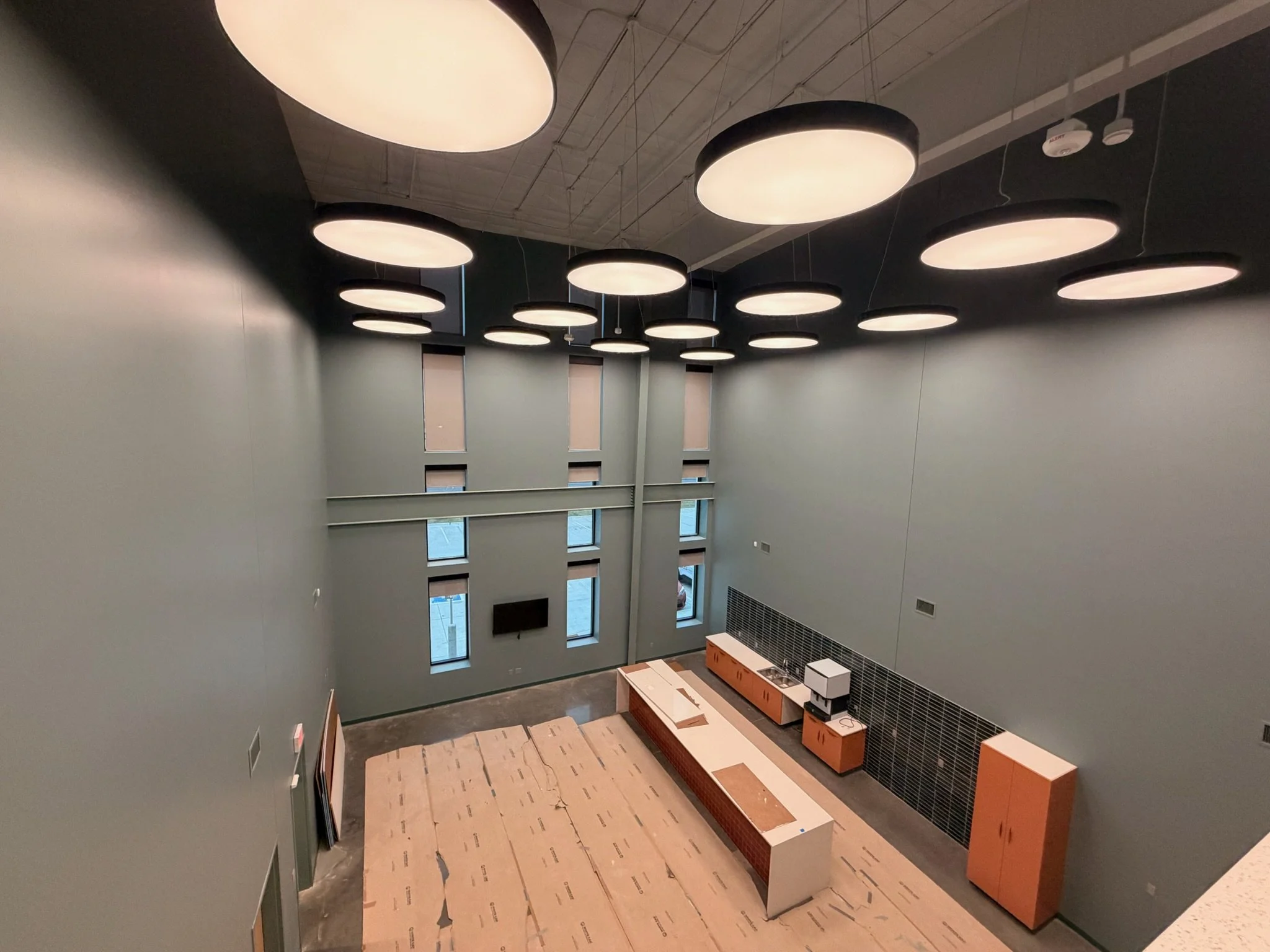 Interior of a modern room with high ceilings, circular light fixtures, multiple small windows, and a kitchen area under construction with cardboard on the floor.