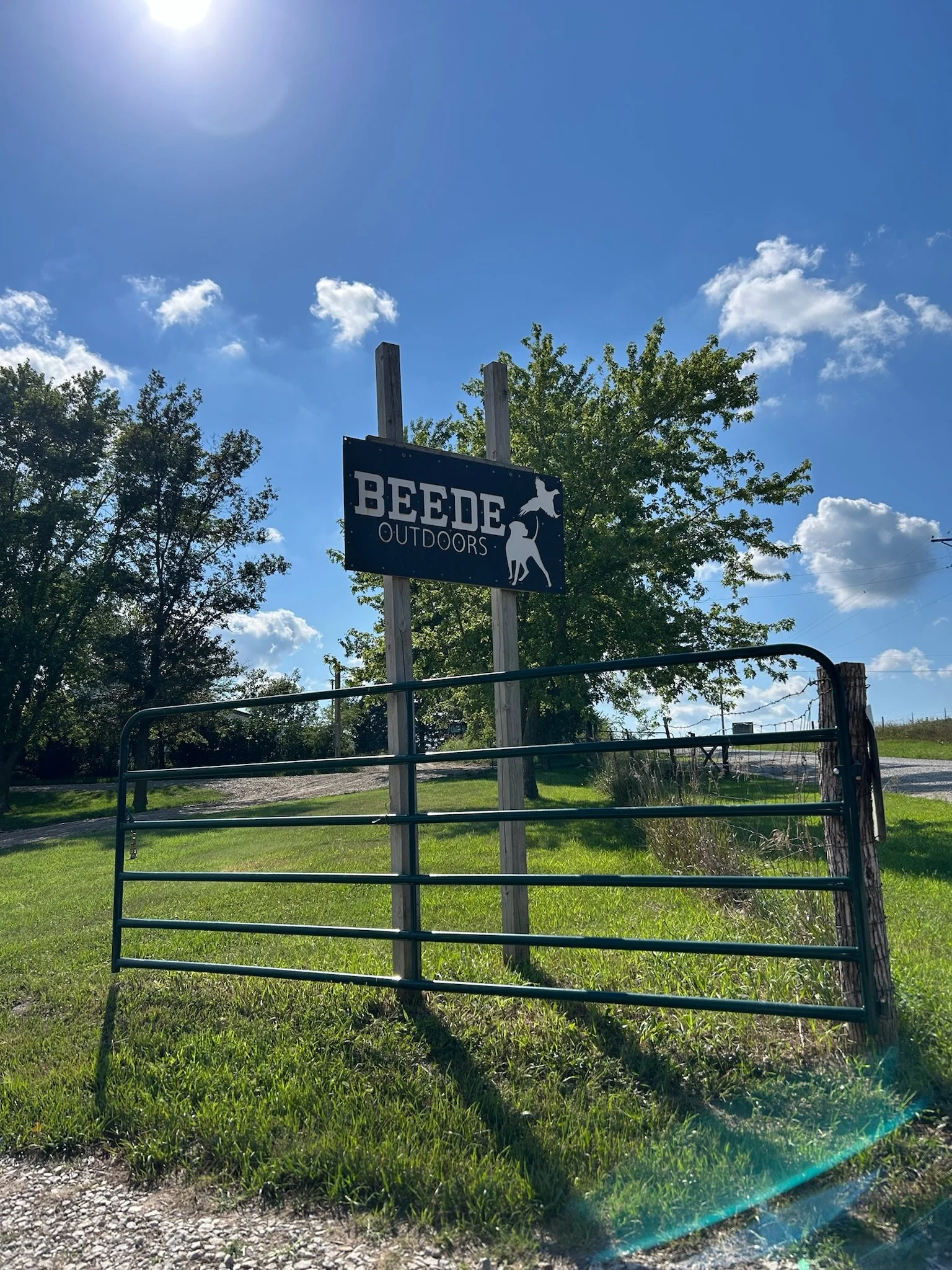 A sign for BEDE Outdoors is mounted on wooden posts, with a blue sky and a few clouds, surrounded by green trees and grass, a metal gate in the foreground, and a gravel path.