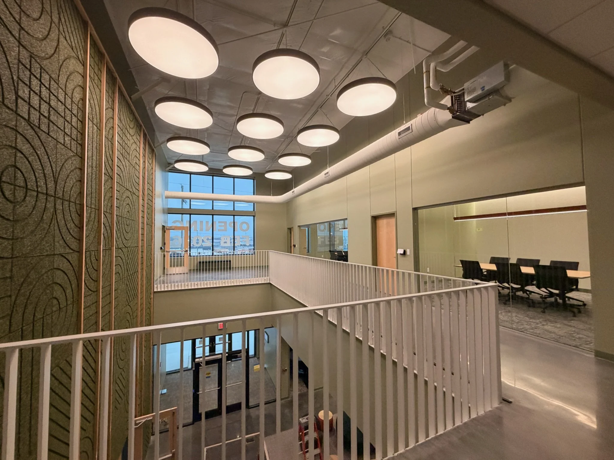 Interior view of a modern building with white circular ceiling lights, an exposed duct system, large windows, and a staircase with a white railing.