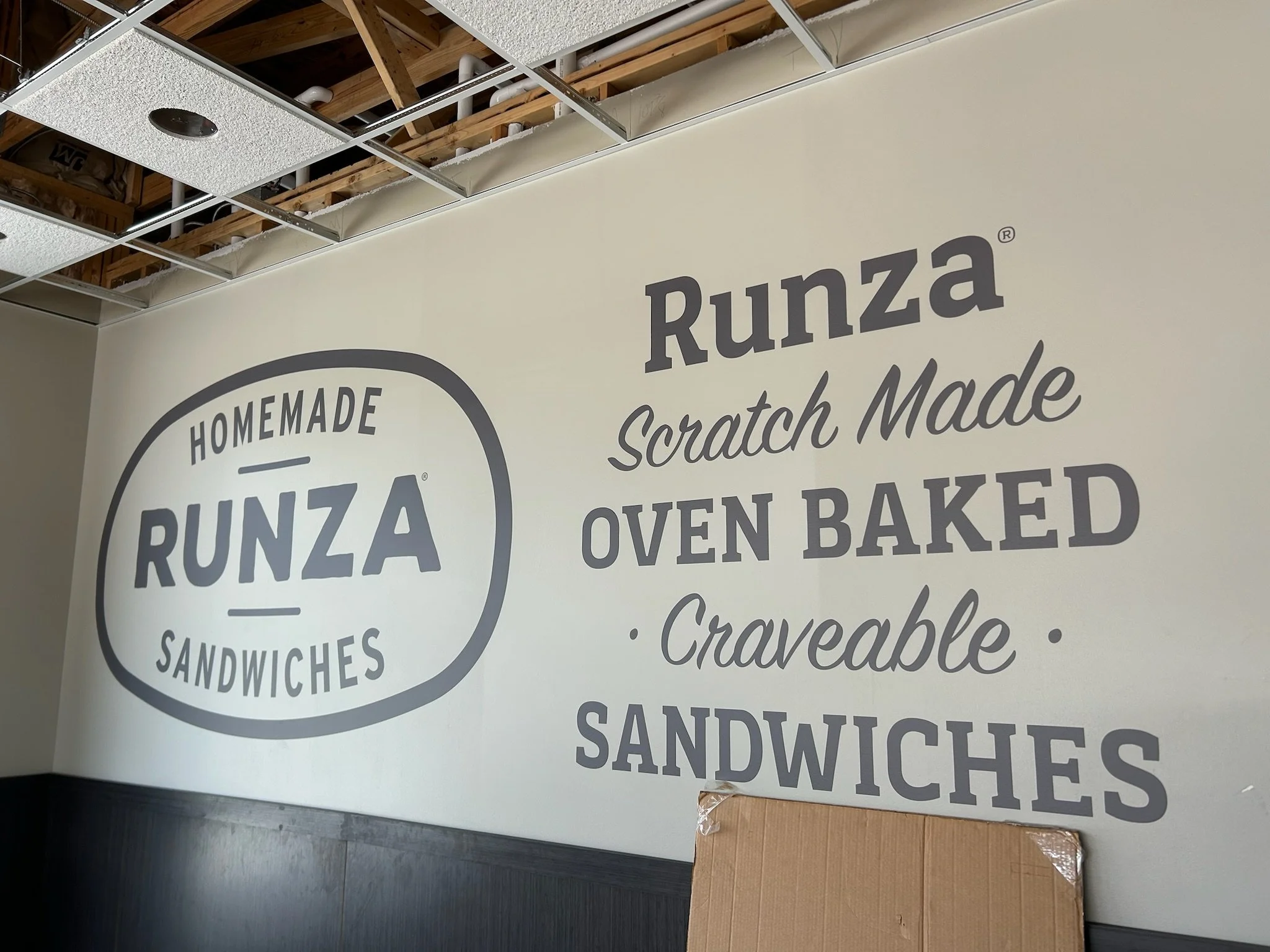 Part of a wall display for Runza, a restaurant offering homemade sandwiches, bread, and cravable baked goods. The display is under construction with exposed ceiling framing and insulation visible.