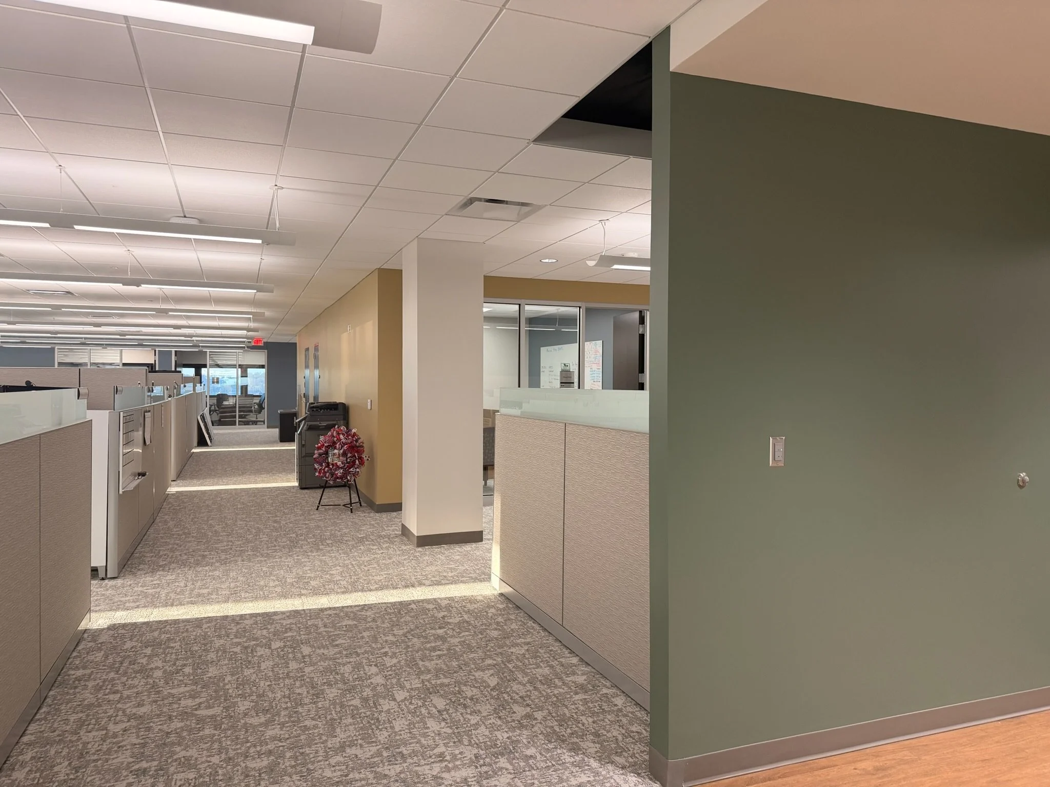 Office corridor with cubicles, beige and green walls, and ceiling with grid tiles and fluorescent lights.