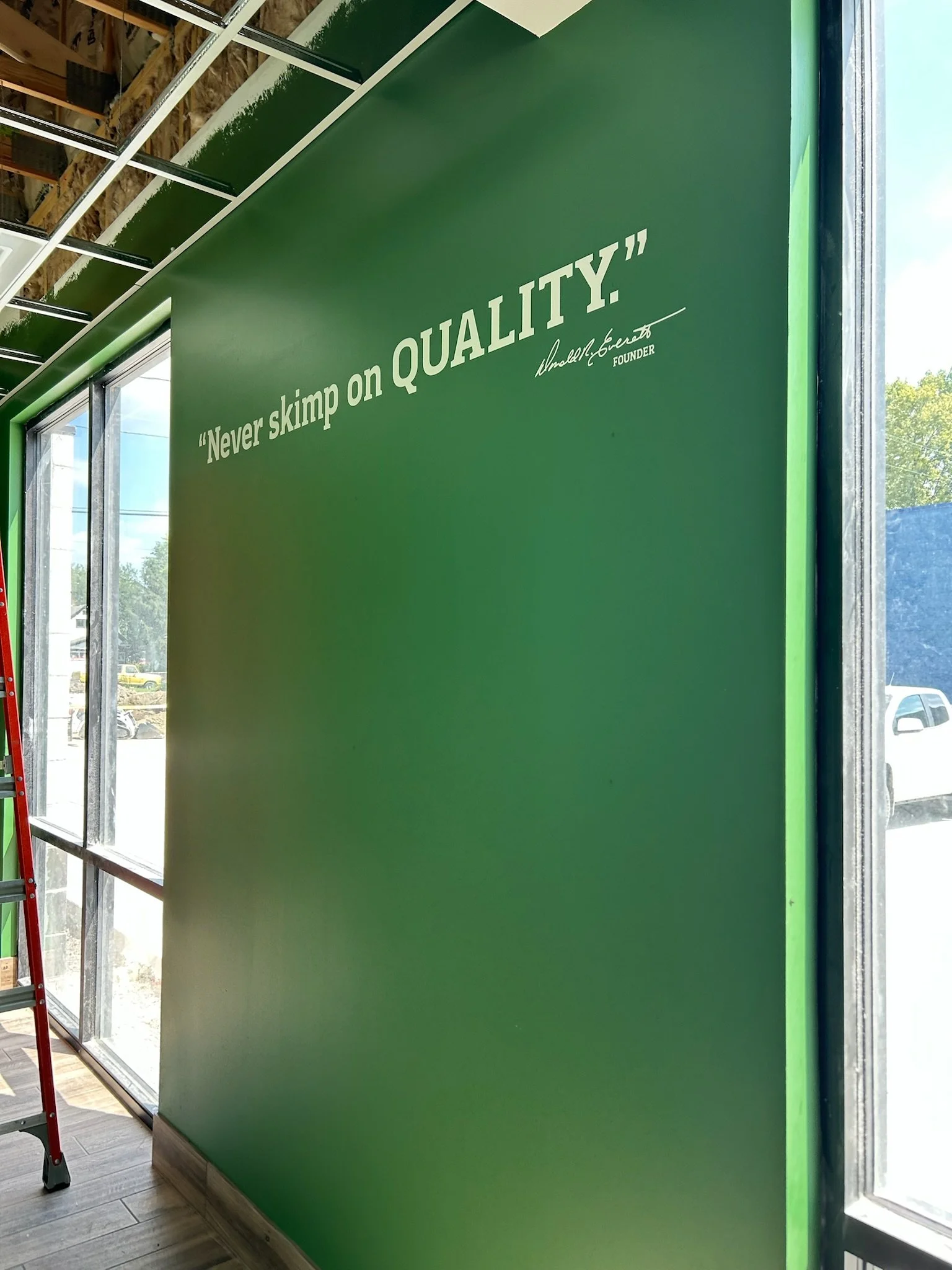 A green wall with white text that reads 'Never skimp on QUALITY' and a signature of the founder, Umarley Convert, below. The wall is inside a building with a large window on the right side, allowing natural light in.