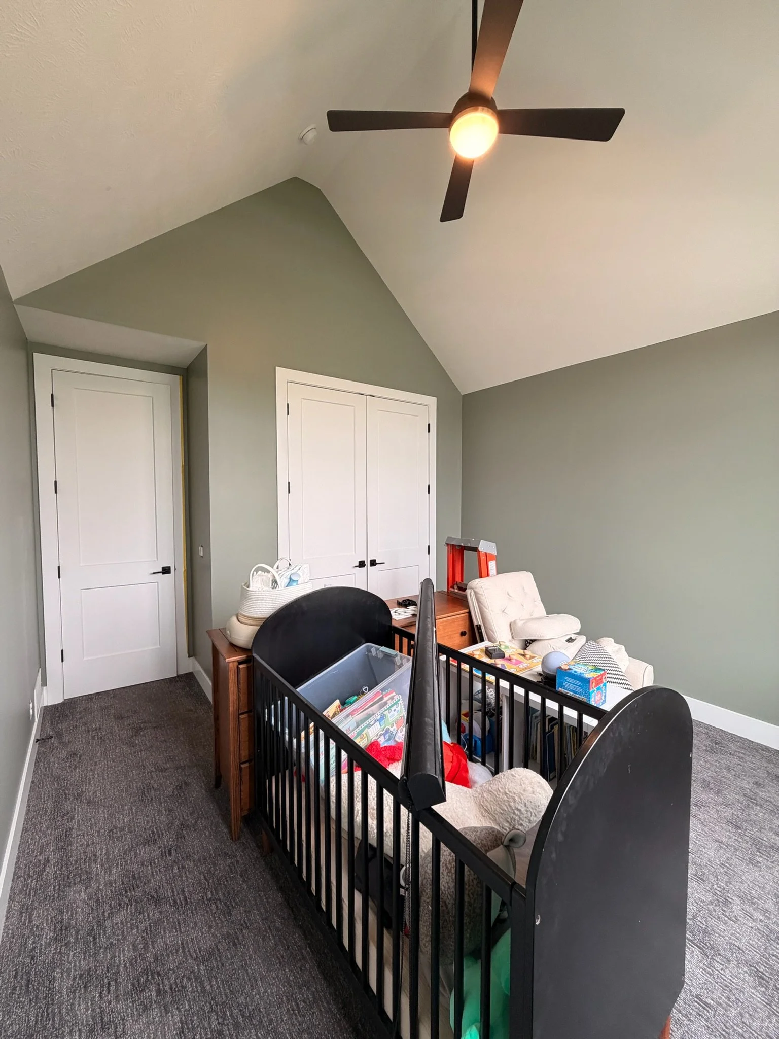 A room with light green walls, two closed white doors, a ceiling fan with light, and furniture including a crib with toys, a small beige armchair, a wooden side table, and various household items.
