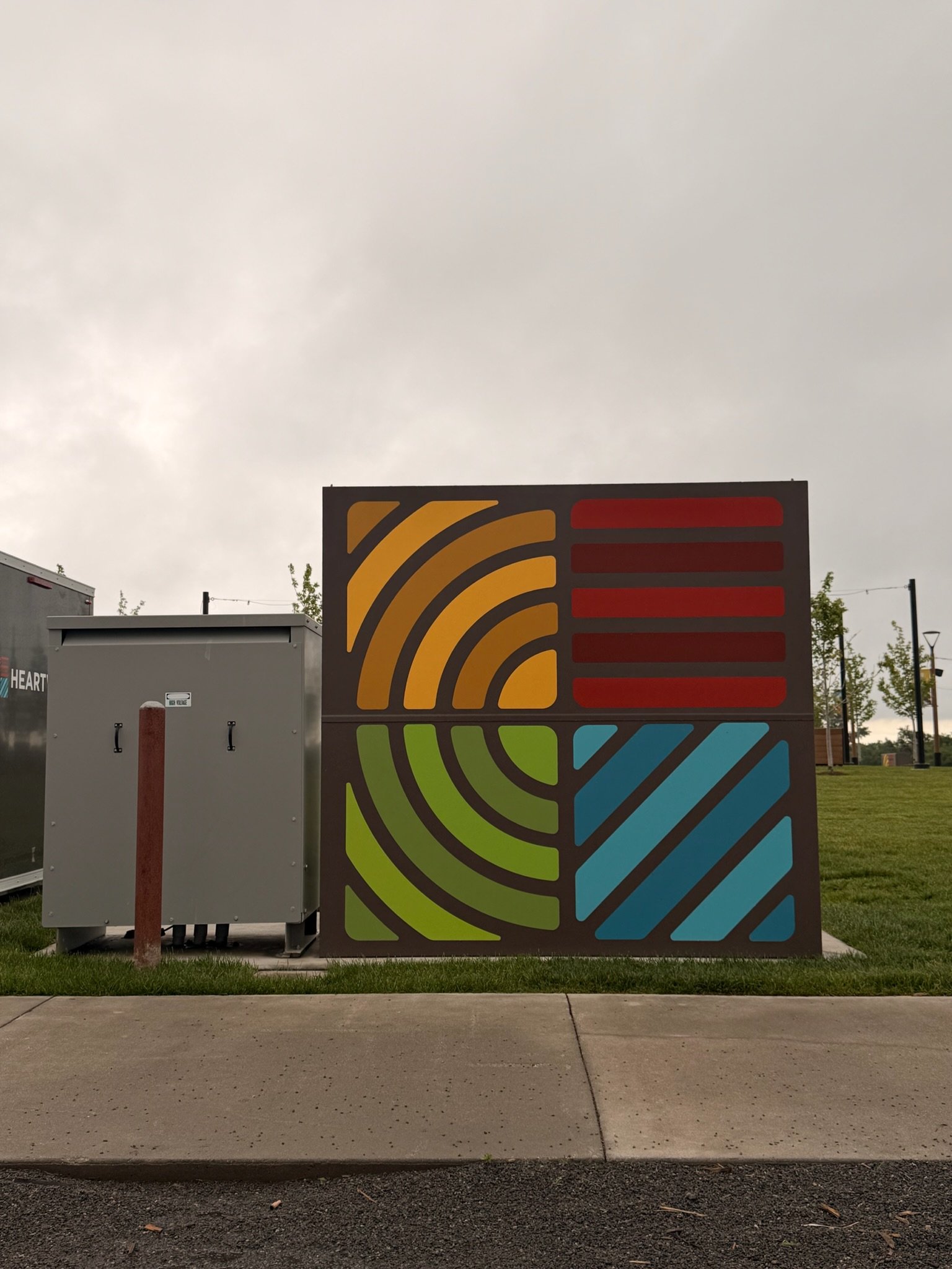 A colorful mural with geometric shapes in yellow, red, blue, and green on a flat outdoor wall, with a cloudy sky above.